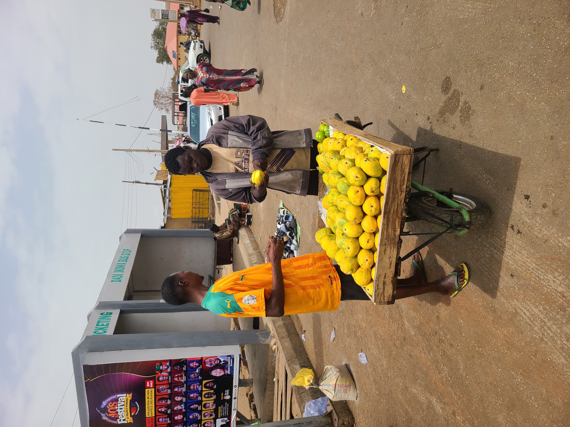 A man is buying mangos from a street vendor in Jos, Nigeria. The vendor stands next to a wooden cart filled with ripe mangos. He is wearing a bright yellow and green soccer jersey. The customer is inspecting a mango. Behind them is a bus stop with an advertisement for the Jos Festival of Praise & Worship, specifically stating that the festival will be held in Philadelphia at #6 Yinga Street in Rayfield, Jos, Plateau State, daily at 3 PM. Other pedestrians and vehicles are visible in the background. The scene is on a paved street with a mix of sunshine and some shadowing.