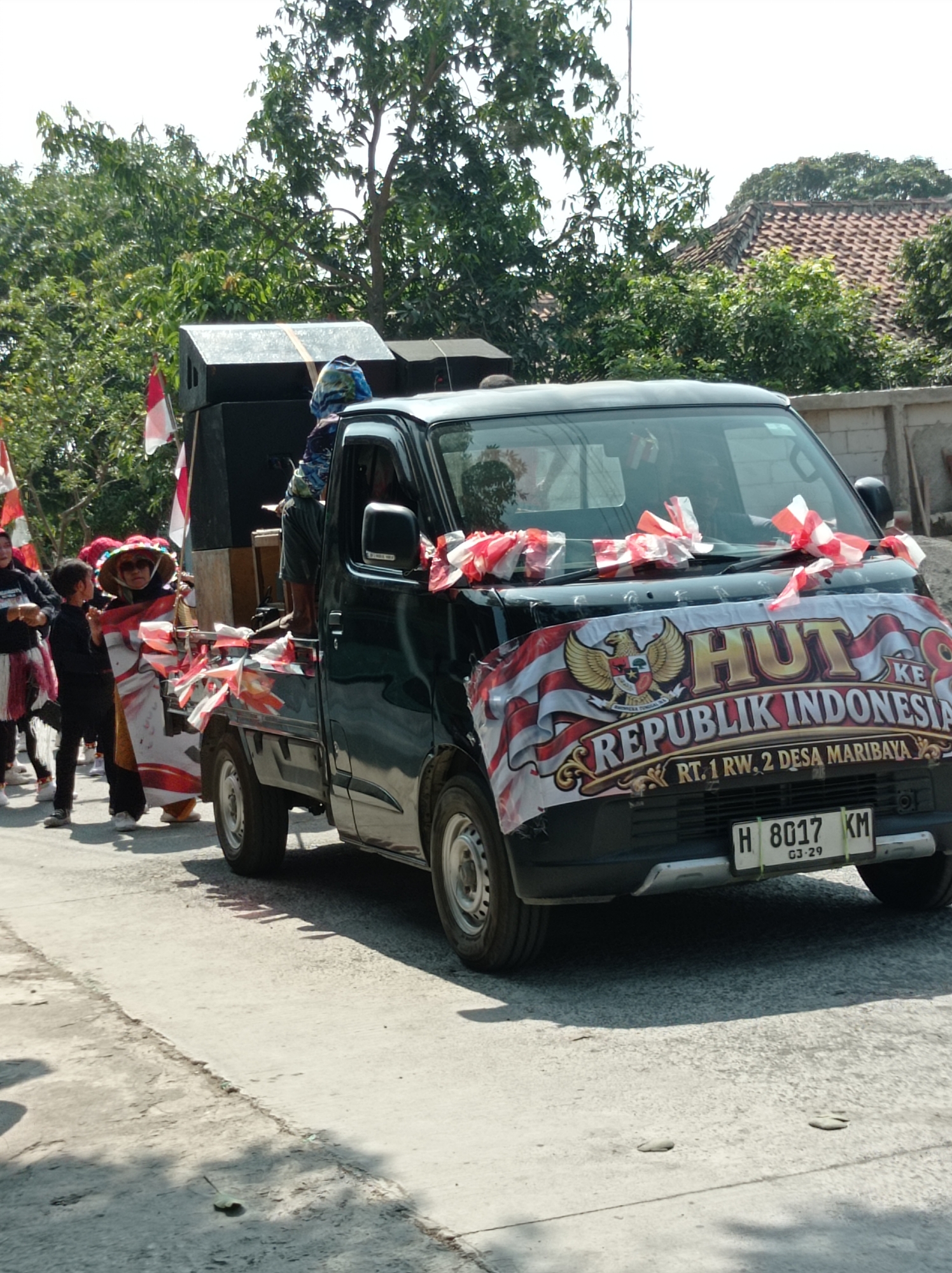 In Maribaya, Indonesia, a dark-colored pickup truck, decorated with a banner that reads "HUT REPUBLIK INDONESIA" and the address "RT. 1 RW. 2 DESA MARIBAYA", is participating in a parade. The banner features an eagle crest. Red and white streamers are tied to the truck's hood and sides. In the bed of the truck, two large speakers are visible, along with a person wearing a patterned blue and white shirt. Several people, including children and adults, are walking alongside the truck. One child, wearing a black outfit, holds a large Indonesian flag. Another person, wearing a conical hat and sunglasses, is dressed in traditional attire adorned with Indonesian flags. The parade is taking place on a paved street under a clear sky, with trees and houses lining the background.
