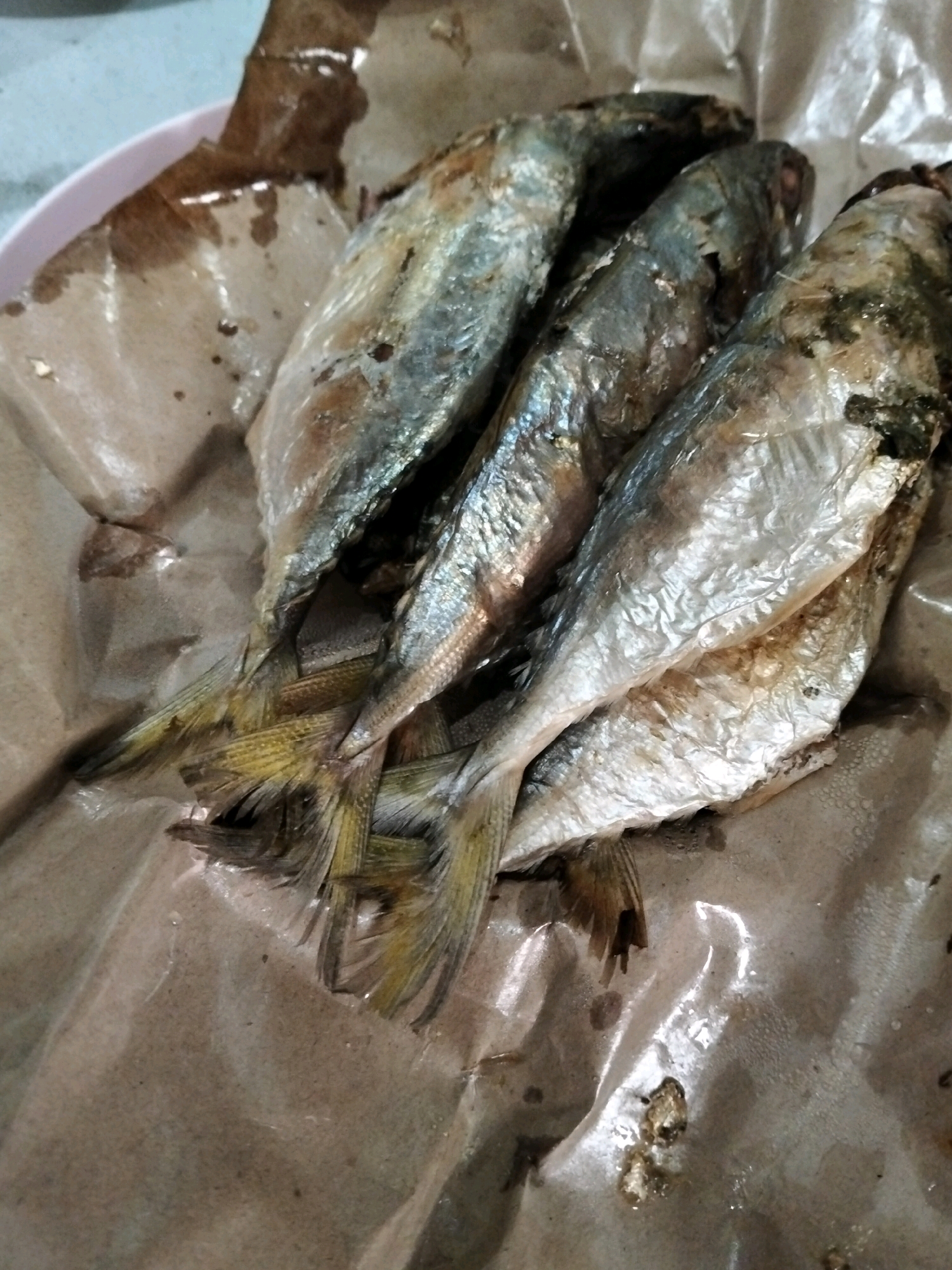 A close-up, overhead shot shows a pile of cooked fish, possibly sardines, served on brown paper. The fish are arranged in a cluster, with their heads facing downwards and tails pointing upwards. Their scales are visible, reflecting light, and some appear to have a slight sheen of oil. The brown paper is wrinkled and shows some watermarks or oil stains. The background is a light-colored surface, possibly a table, with some blurred out elements. The scene appears to be in a casual setting, possibly a street food stall or a home kitchen in Central Jakarta, Indonesia.