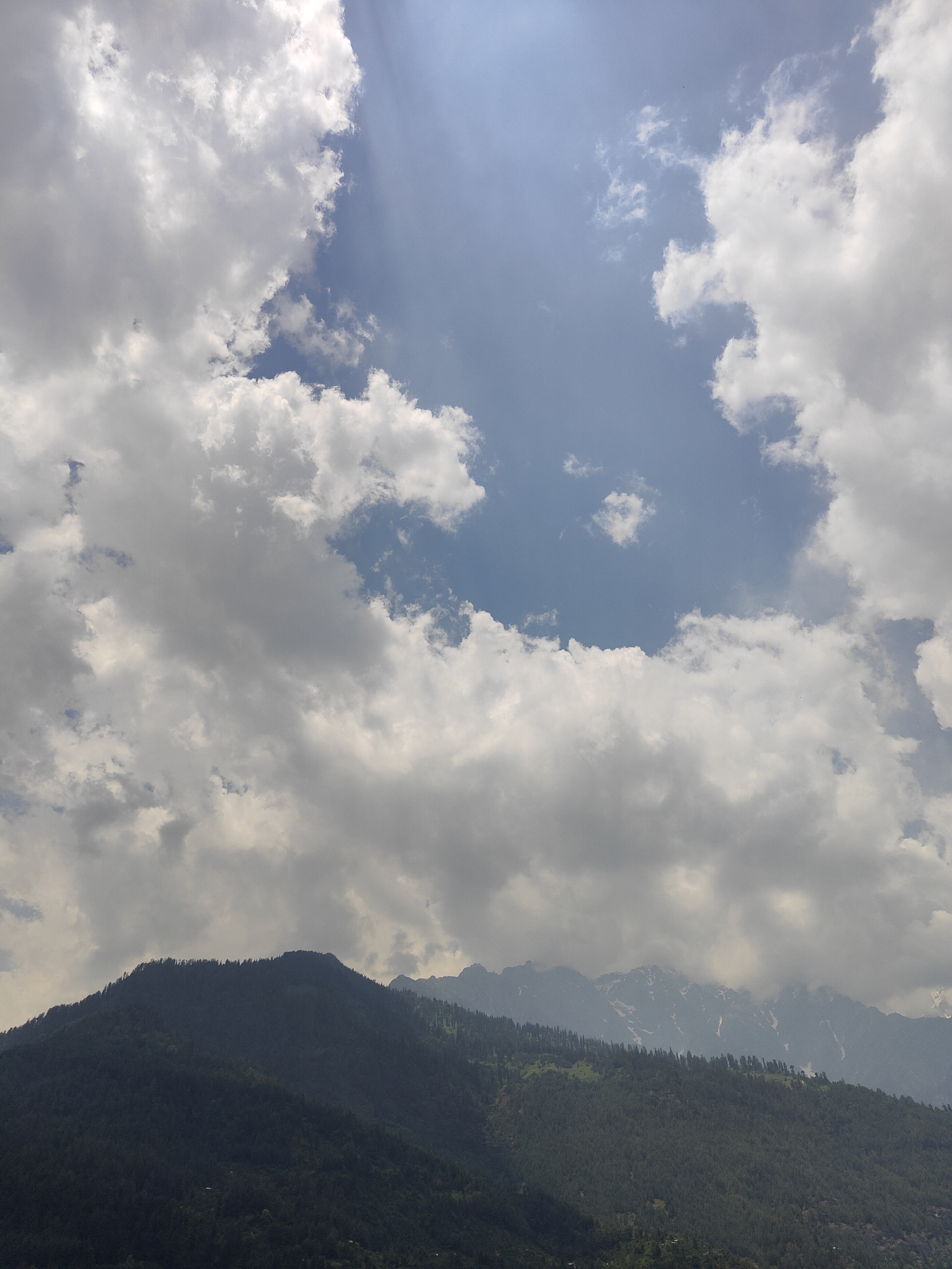 A vertical, medium shot photograph captures a mountain landscape under a cloudy sky. The upper half of the image is dominated by a dynamic sky filled with white and grey clouds, with patches of blue sky visible. Sunlight streams through a gap in the clouds, creating a bright, ethereal effect. The lower half of the image features the dark, tree-covered slopes of mountains. The trees form a dense canopy, obscuring most of the terrain. In the distance, the jagged peaks of further mountains are visible, some with patches of snow. The overall impression is of a natural, mountainous environment. The photograph was taken in Vashisht, India.