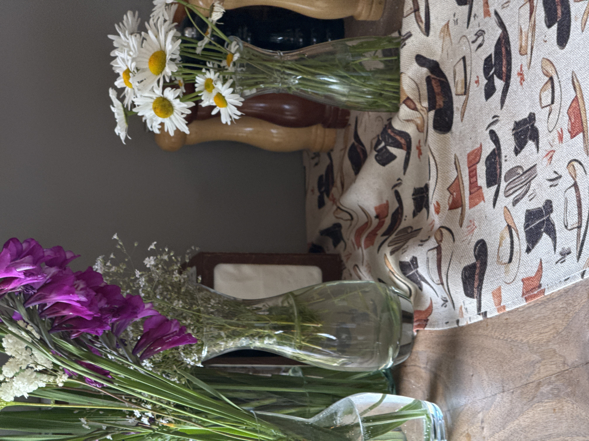 This is a close-up, eye-level, indoor shot of a rustic table setting. In the foreground, a light-colored fabric runner is spread across a wooden surface. The runner is adorned with a repeating pattern of cowboy hats, boots, and cacti in shades of brown, black, and orange.
To the left, a clear glass vase holds a bouquet of tall, thin green foliage and delicate white flowers. Beside it, another clear glass vase, wider at the base and narrowing towards the neck, contains a spray of vibrant purple gladiolus flowers and more small white filler flowers.
To the right of the purple flowers, a vase with a similar shape to the one holding the gladiolus contains a bouquet of white daisies with yellow centers. Behind the daisy bouquet, three wooden salt and pepper grinders are visible. The grinder on the left is a lighter wood, the middle one is dark brown, and the one on the right is a darker shade of brown or black. A dark, rectangular object, possibly a napkin holder with a decorative wood frame, stands behind the purple flowers.
The background is a plain, dark gray wall. The overall scene evokes a western or country aesthetic. There are no people visible in the image. The lighting suggests an indoor setting, possibly with natural light coming from the left, casting subtle shadows.
While the image depicts a table setting, its specific location within the city of Dsegh, Armenia, cannot be determined from the visual information provided.