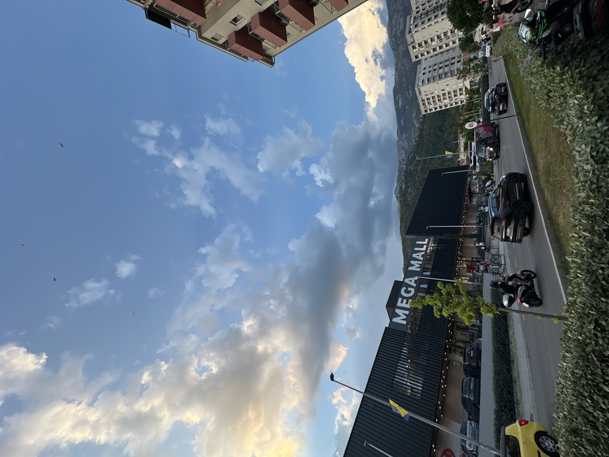 This is a slightly tilted, eye-level shot of a street scene in Budva, Montenegro, during the late afternoon or early evening. The sky is a mix of blue with scattered clouds, some of which are illuminated with a warm, golden light, suggesting sunset. A large, modern building with the prominent text "MEGA MALL" is visible on the left. The mall has a dark facade and appears to be open for business, with lights on inside.
In front of the mall, a paved road runs parallel to it. Several cars are parked along the side of the road, and a few vehicles are driving. A motorcycle with two riders is passing by on the road. To the right of the road, there is a dense green hedge. In the background, tall apartment buildings and a steep, tree-covered mountain range are visible, indicating the geographical setting of the town. The overall mood of the image is calm and suggests a typical day in a city with both commercial activity and natural beauty.