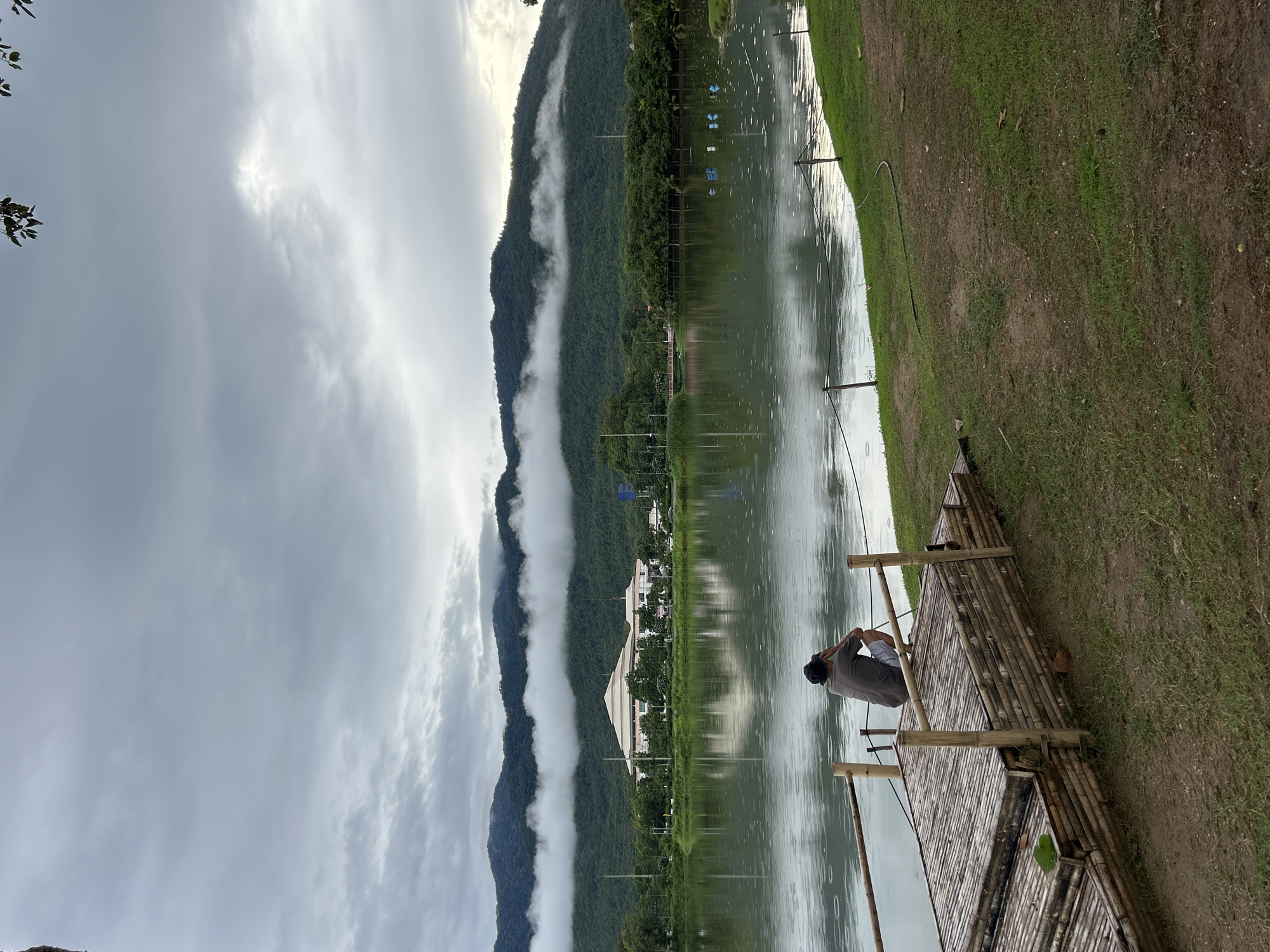 A person is crouched on a bamboo raft by the water, facing away from the camera. The raft is made of tightly packed bamboo poles and is moored to a wooden railing. The person is wearing a dark t-shirt and light-colored shorts, with a dark hat. They appear to be looking out at the lake and the surrounding landscape.
The lake is calm, with ripples on the surface indicating light rain. In the background, across the lake, is a large, modern building with a distinctive white roof, surrounded by lush green trees. Beyond the building, verdant hills rise up, partially obscured by a band of white clouds that hug their slopes. The sky is overcast with heavy, grey clouds. The foreground shows a grassy bank with some dirt paths. The overall atmosphere is serene and perhaps a little melancholic due to the weather. The scene is likely at a park or recreational area, possibly the Mae Hia Agricultural College lake in Chiang Mai, Thailand, given the building style and the natural surroundings.