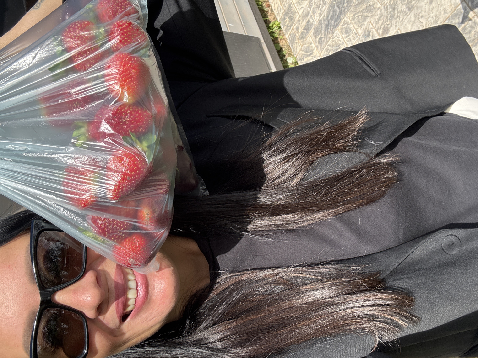 A woman wearing sunglasses and a black blazer holds a clear plastic bag filled with strawberries. Her mouth is slightly open, revealing her teeth. The background shows a paved surface with some greenery. The image is taken outdoors on a sunny day. No specific location in the city of Chaikovsky, Russia is identifiable.