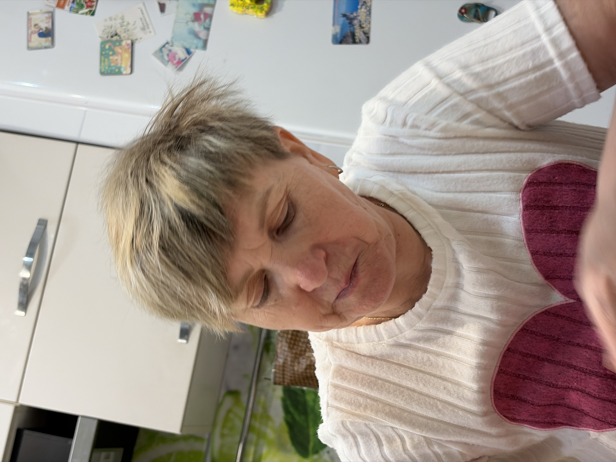 A close-up shot reveals an older woman, positioned indoors, partially obscured by a light. Her hair is short, light brown with some blonde highlights, and appears slightly tousled. Her eyes are closed or cast downwards, suggesting a moment of reflection or fatigue. She is wearing a light-colored, ribbed shirt with decorative heart-shaped patches. A thin gold necklace is visible around her neck.
Behind her, partially visible, is what appears to be a kitchen setting. A white cabinet is noticeable, with a silver handle. Above the cabinet, there are multiple small magnets or pictures affixed to a surface, possibly a refrigerator. The background lacks further detail to allow for a definitive identification of the location in the city of Noviy, Russia.