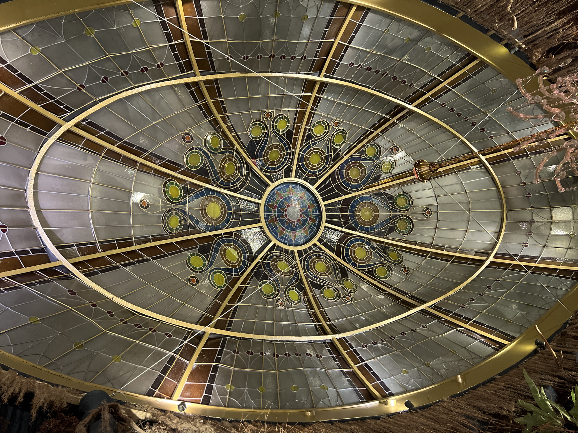 This media file captures an upward view of an elaborate, oval-shaped stained glass ceiling or skylight. The design features a complex interplay of geometric and organic patterns, primarily in a style suggesting Art Nouveau or Art Deco influences.
The stained glass consists of numerous panels held within a golden-colored metal framework. The outer sections are dominated by translucent gray, white, and brown-amber glass segments, arranged in a grid-like pattern with occasional small red or yellow-green circular accents. As the design approaches the center, more vibrant colors emerge. Radial patterns, reminiscent of stylized peacock feathers or eyes, are formed by concentric yellow-green and blue circles, often outlined in gold, against darker glass. The very center of the oval displays a symmetrical star or flower motif rendered in a variety of colors including blue, red, and earthy tones.
A thick, golden-colored oval frame meticulously outlines the entire stained glass structure. Beyond this frame, the surrounding ceiling or wall is covered with a dark, heavily textured material, appearing to be composed of natural fibers or woven straw, which contrasts with the smooth glass and metal.
In the upper right corner, a portion of a decorative light fixture or chandelier is visible. It features clear, pinkish glass elements shaped like delicate branches or flowers, supported by a golden metallic rod. Several thin white wires can be seen running across different sections of the stained glass, possibly for lighting or structural purposes.
No people are present, and no specific activity is depicted. The image focuses on the intricate architectural details and decorative artistry, suggesting an indoor setting in a finely crafted building within the Ubud District, Indonesia. No visible text is present.
