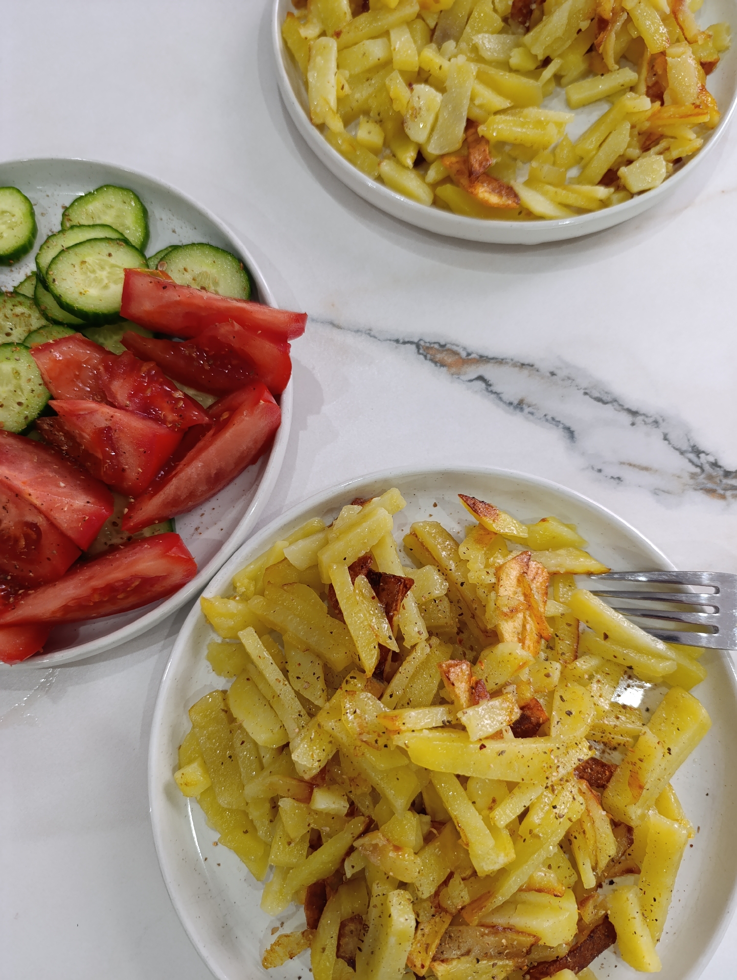 The image depicts a table with three plates of food. One plate contains sliced cucumbers and tomatoes seasoned with herbs or spices. The other two plates hold fried potatoes, also seasoned. A fork rests on one of the potato plates. The setting appears to be indoors, likely a home kitchen or dining area, with a white marble or similar countertop. Based on prior context, the location is Chaikovsky, Russia, but this is not apparent from the visual information in the image. No people are visible.