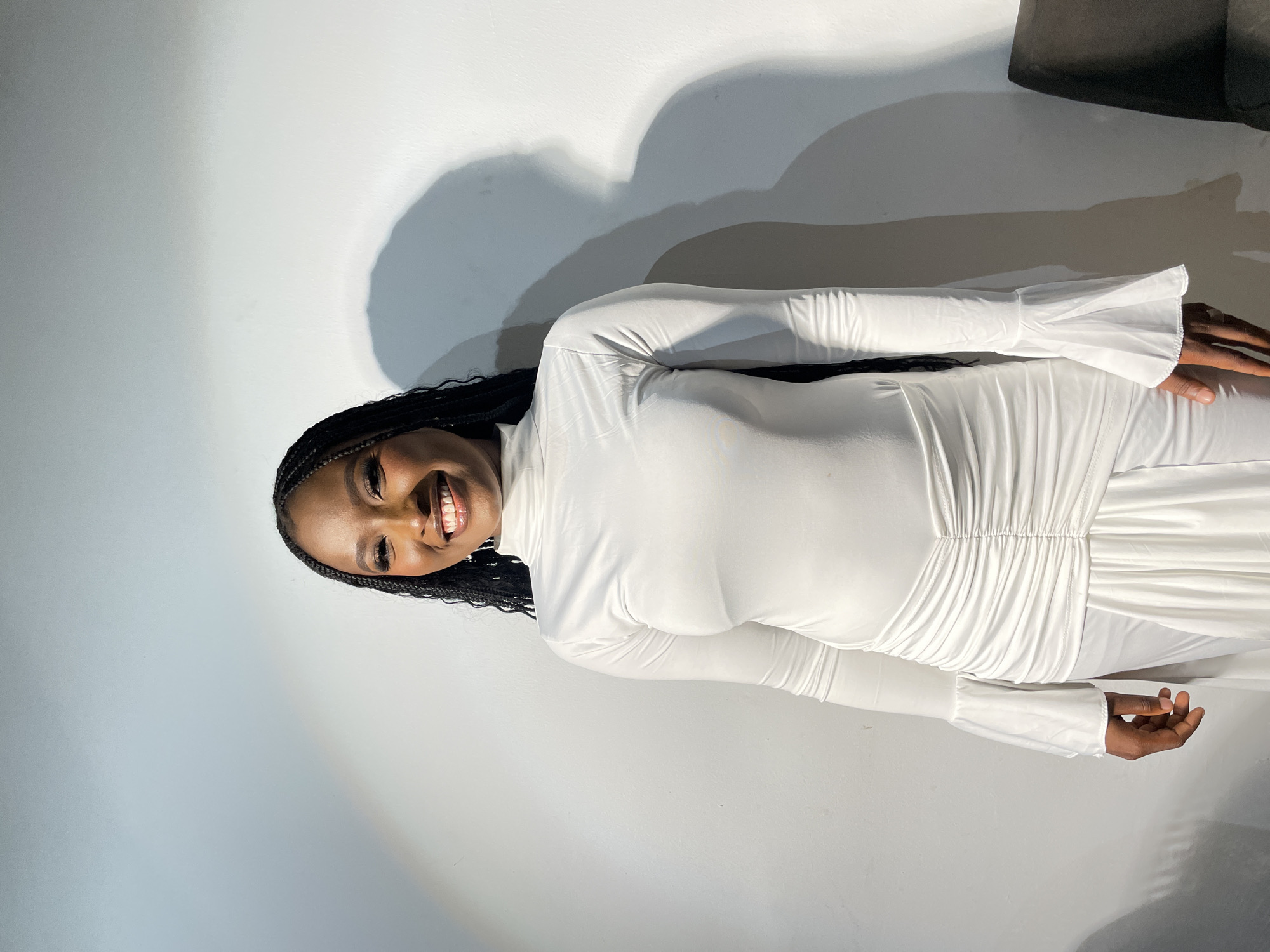 A woman with braided hair is wearing a long-sleeved white dress. She is smiling and facing the camera. She is standing against a white wall with shadows. The location is most likely a studio or indoor space in Barakin Akawo, Nigeria.