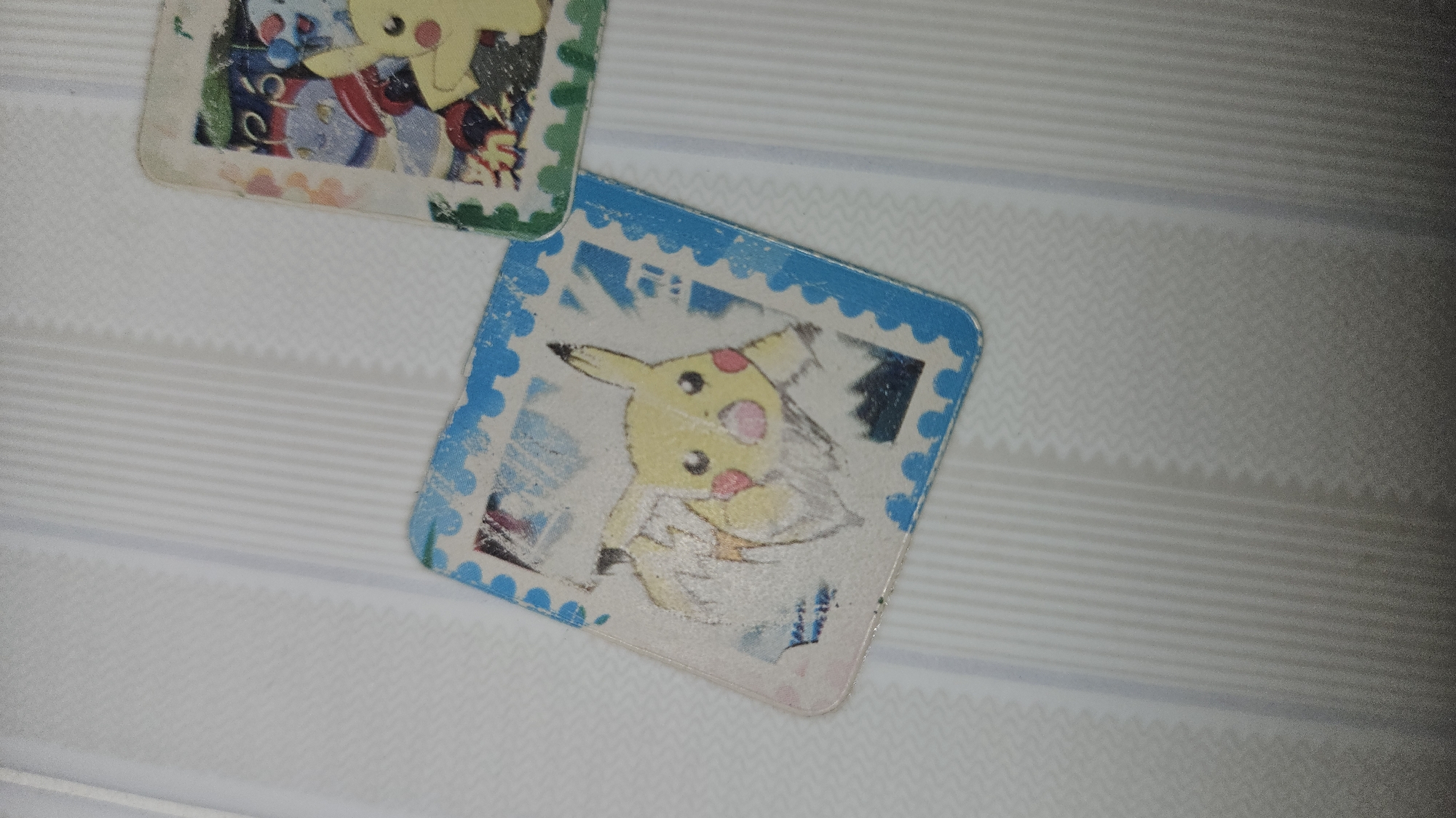 The image shows two Pokemon Pogs lying on a white surface with a subtle pattern. The pogs feature Pikachu, a popular Pokemon character. The pog in the foreground has a blue, stamp-like border, while the other has a green border. They are likely located in Cam Đức, Vietnam.