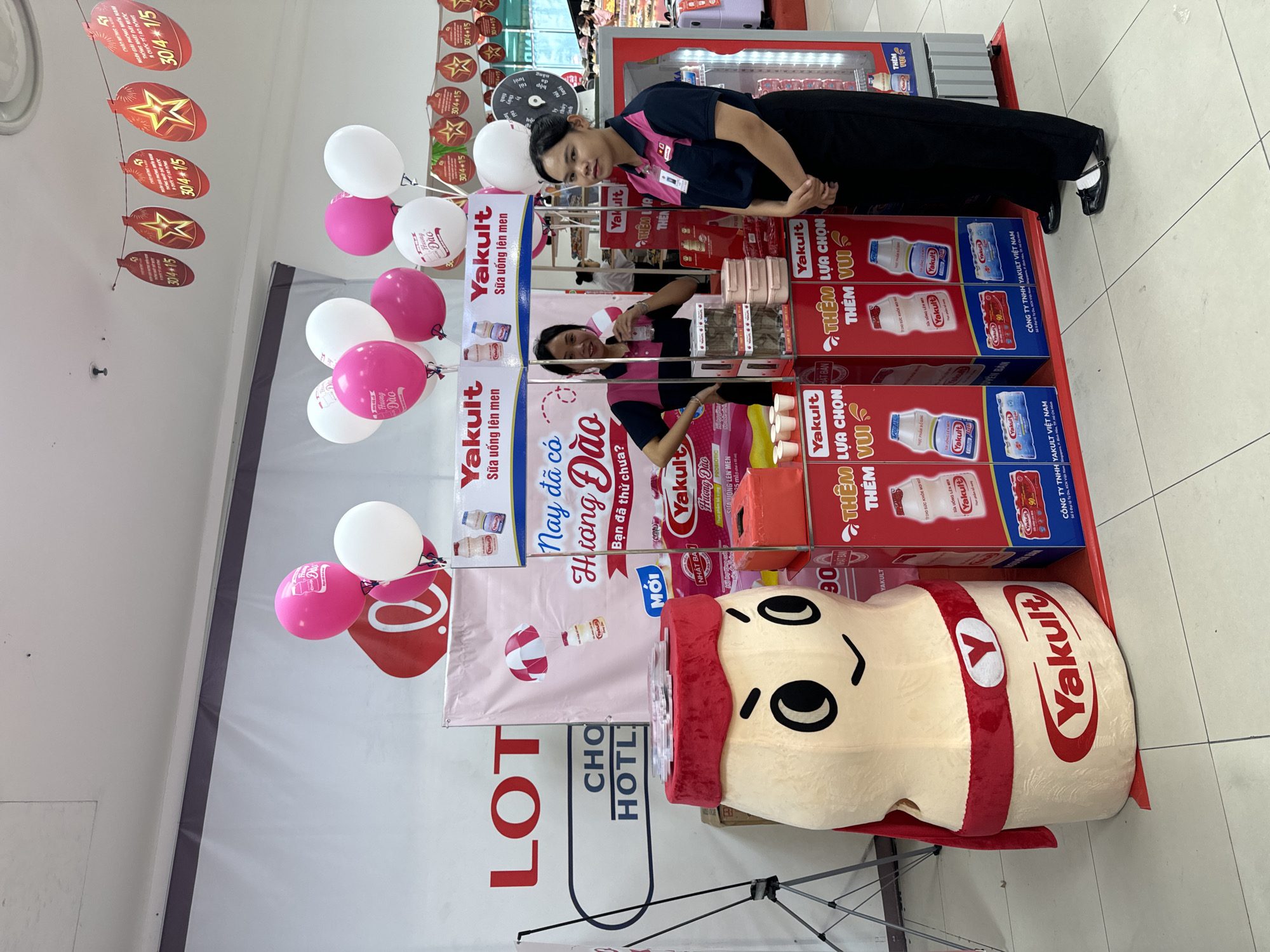 Captured in Phú Thủy, Vietnam, the scene unfolds inside a LOTTE Mart, where a woman stands beside a promotional display for Yakult. The display is composed of Yakult product boxes and advertising materials, including posters featuring the text "Yakult", "Sữa uống lên men", and "Nay đã có Hương Đào". The woman, dressed in a dark top with pink accents and dark pants, stands near a large Yakult mascot. Pink and white balloons are attached to the display. Strings of promotional tags hanging from the ceiling add to the scene.
