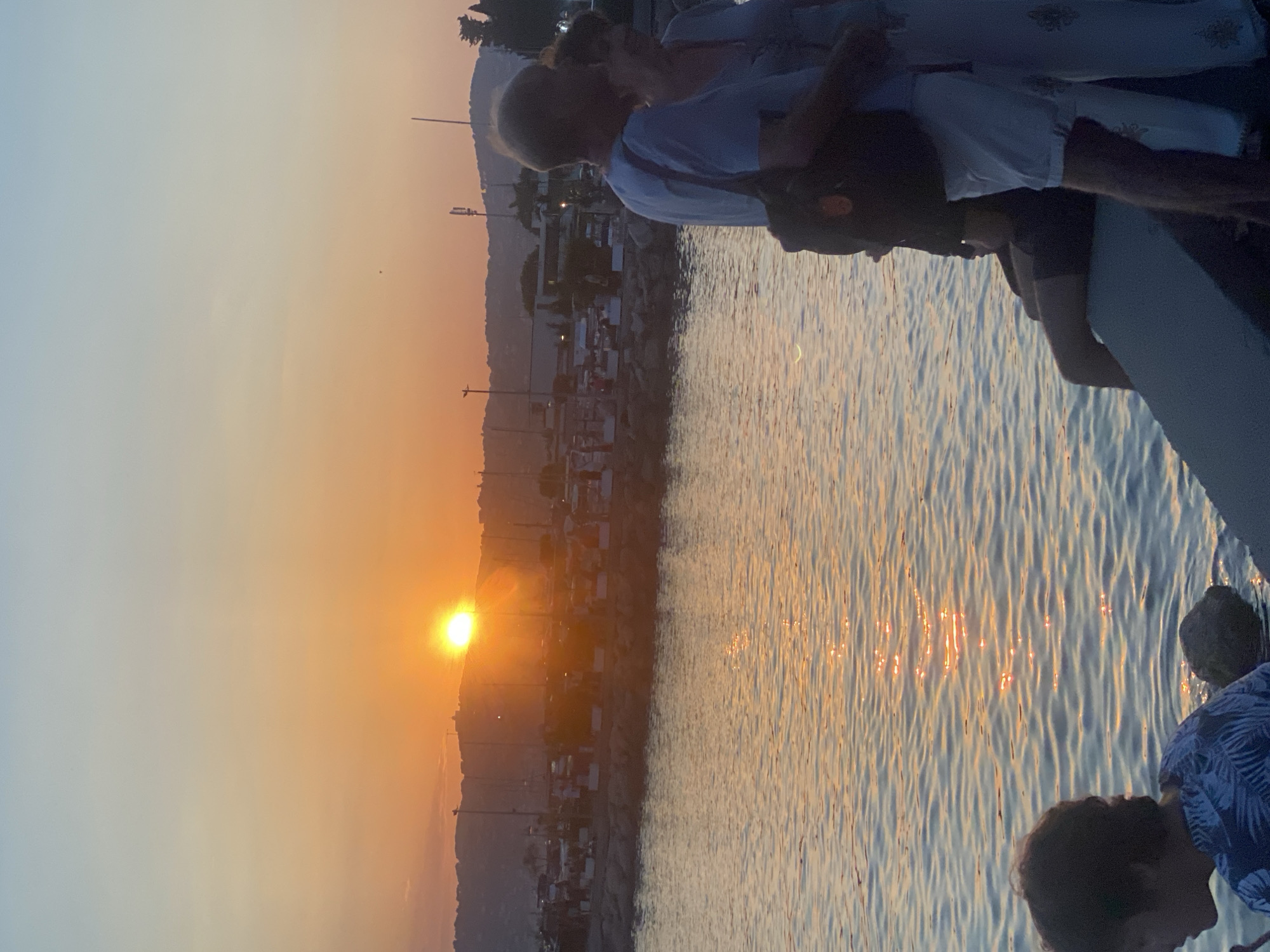 The media file captures a sunset scene in Antibes, France. The image is oriented vertically, with the sun low on the horizon, casting a warm, orange glow across the sky and reflecting on the calm water. In the foreground, several people are visible, some of whom appear to be looking out at the sunset. One person in the lower right corner wears a patterned shirt. The middle ground shows a coastline with hills and buildings, possibly a town or resort, with lights beginning to illuminate. A marina with boats is also visible along the coast. The water is rippling gently, creating shimmering reflections of the sunset. The overall mood is peaceful and serene, characteristic of a sunset by the sea. The time of day is clearly late evening, as indicated by the setting sun and the quality of light. The weather appears to be clear and warm.