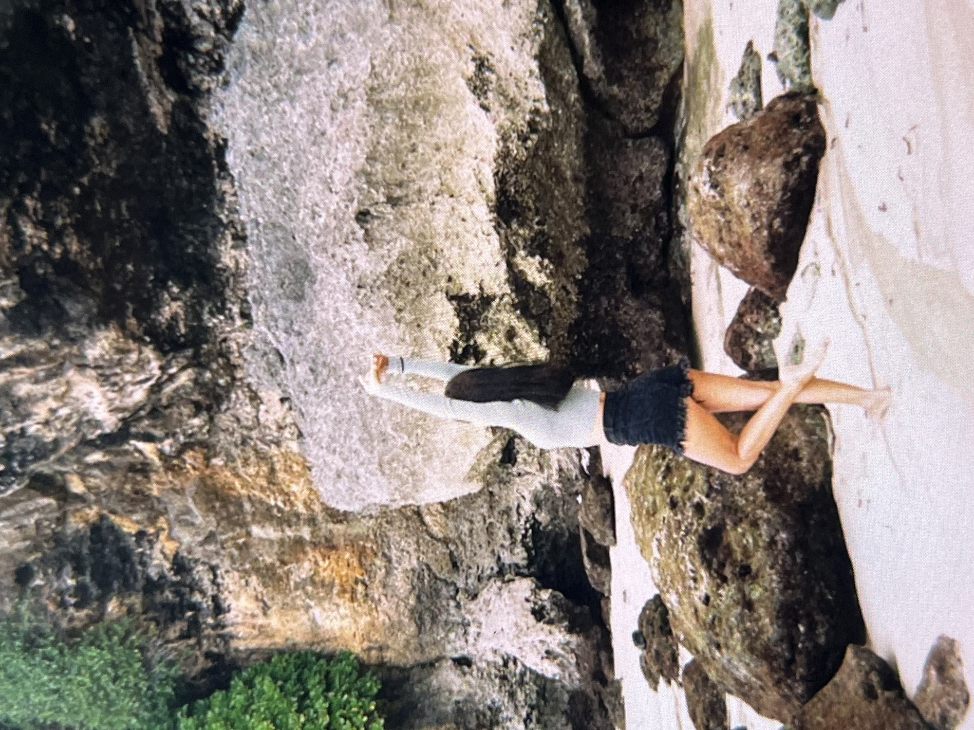 A young woman with long, dark hair is positioned in a prone position atop a large, moss-covered rock formation. She wears a light-colored, long-sleeved shirt and dark denim shorts with frayed edges. Her legs are extended and crossed at the ankles, with her bare feet slightly off the edge of the rock.
The rock formation is situated on a sandy beach, adjacent to a cliff face. The cliff is textured and layered with vegetation growing at its base. A waterfall cascades down the cliff face.
The location is Unknown, Indonesia.