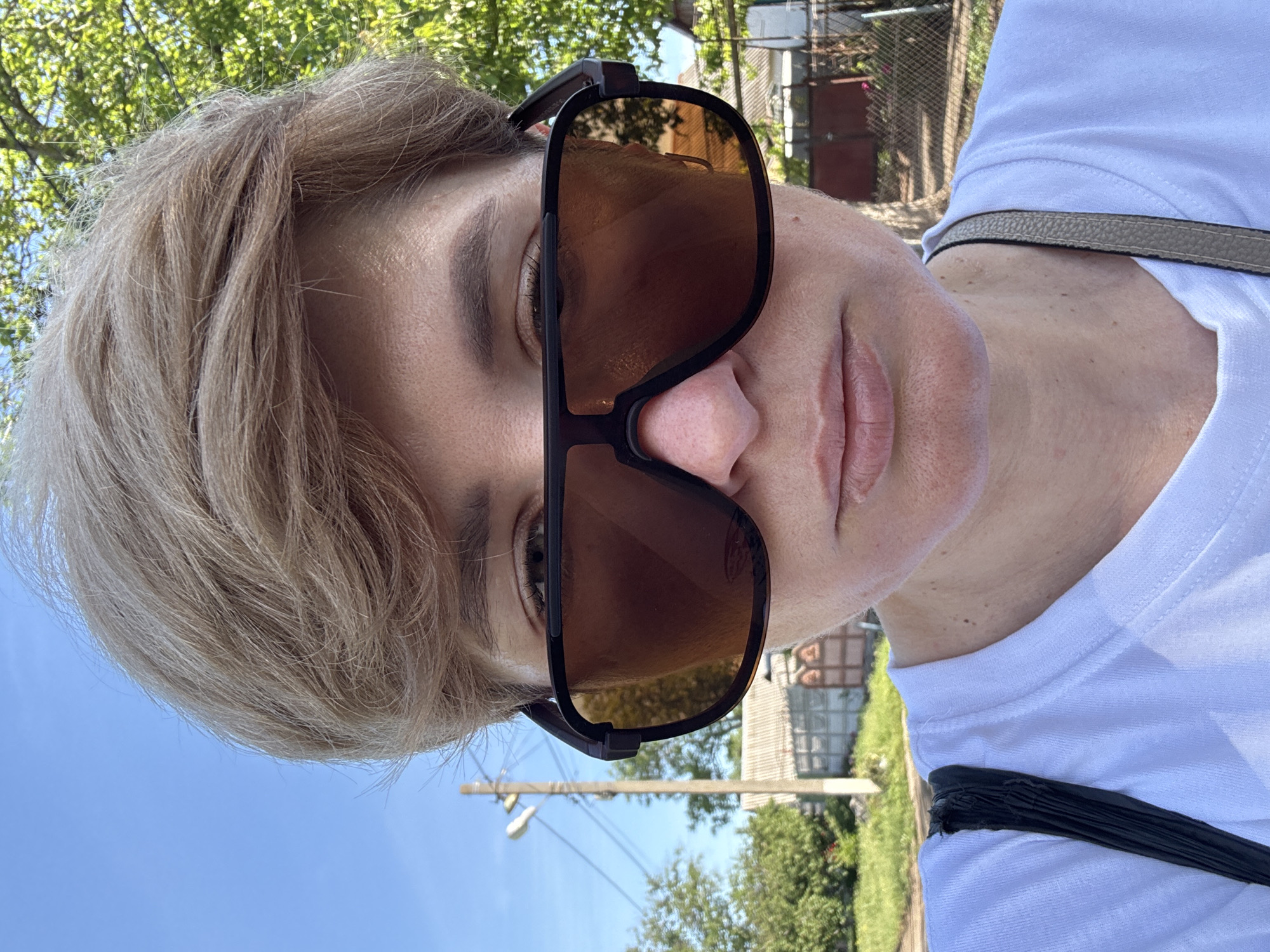 A close-up selfie captures a person from a low angle, looking upwards towards the camera, their head tilted back. The individual has short, layered, light-colored hair, appearing blonde or light brown with a somewhat tousled texture. They are wearing large, dark-framed sunglasses with brown tinted lenses, which cover most of their eyes. Part of their right eye is visible beneath the top rim of the sunglasses, showing a light-colored iris.
The person has visible pores on their skin, with some small freckles or moles on their neck and face. They are wearing a plain white t-shirt. A narrow, light grey or tan strap from a bag or purse is visible over their right shoulder, contrasting with the white fabric. Another black strap, possibly from the same bag, is seen lower on their torso.
The background, visible around and reflected in the sunglasses, indicates an outdoor setting on a clear, sunny day. A bright blue sky is prominent at the top and left of the frame. Green trees with lush foliage are visible, suggesting a natural environment. In the mid-ground and right side, there are hints of a residential or rural area, including power lines, a utility pole, and parts of distant buildings or fences, bathed in sunlight. There is no visible text in the image.