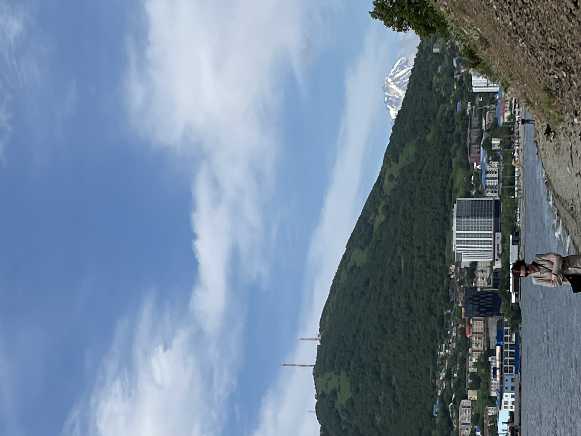 This is a vertically oriented, eye-level, medium-distance photograph taken on a sunny day in Petropavlovsk-Kamchatsky, Russia. In the foreground, a person wearing sunglasses and a trench coat stands with their arms crossed, facing slightly left. They are on a rocky shoreline next to a body of water.
Behind the person, a city skyline is visible. Several buildings of varying heights and architectural styles are present, including a modern, tall, white building with many windows. In the middle ground, a large, densely forested green hill rises. Two tall, red and white striped radio towers stand on top of the hill.
In the background, a snow-capped volcano is visible against a partly cloudy blue sky. The volcano appears to be smoking slightly. The overall scene depicts a coastal city nestled against a dramatic natural landscape of hills and a volcano.