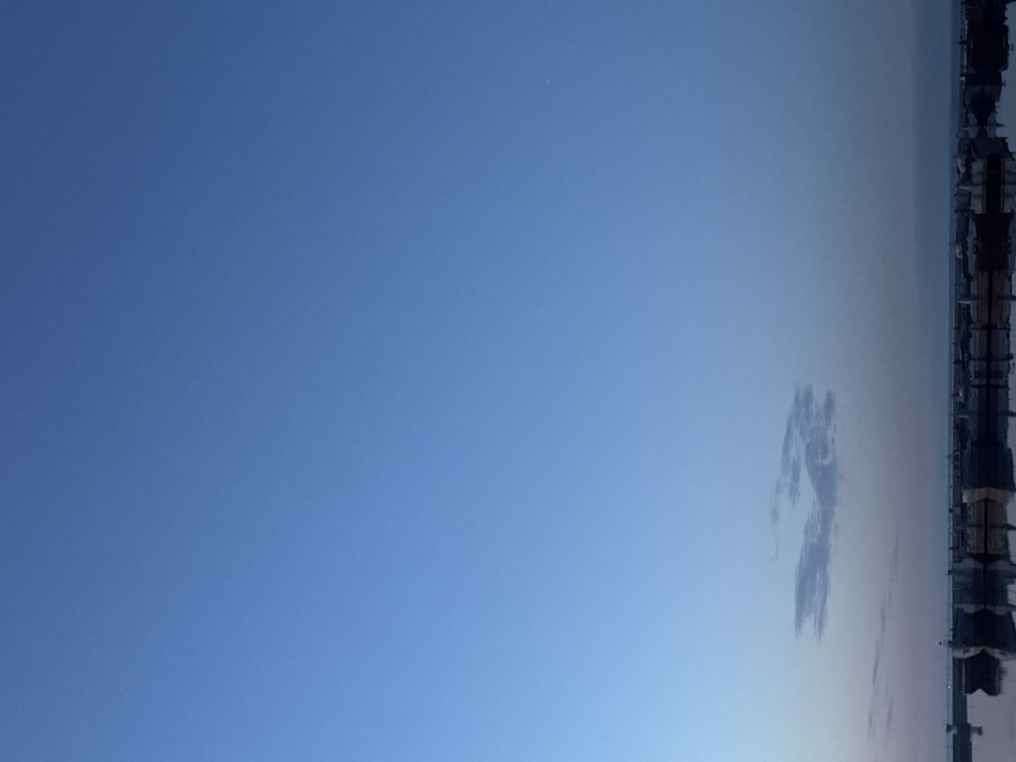 The image displays a serene outdoor scene dominated by a vast, clear blue sky, rotated 90 degrees clockwise in the provided original. When viewed correctly, the sky occupies the majority of the frame, showcasing a gradient from a deeper blue at the top to a lighter, paler blue-grey closer to the horizon. A very small, faint white point, possibly a star, distant planet, or aircraft, is visible in the upper right portion of the blue sky.
Along the distant horizon, a dark, complex silhouette of what appears to be a cityscape or urban structures is visible. These structures, likely buildings with varying rooflines and vertical elements, are indistinct due to distance and lighting conditions. Below these structures, especially in the larger crops, there's a suggestion of a reflective surface, possibly water, mirroring the lower parts of the distant scene. A few light, wispy, elongated clouds are scattered near the horizon, just above the silhouetted buildings.
The lighting is soft, suggesting either early morning or late afternoon, as the distant structures are mostly in silhouette. There are no people or specific activities depicted. The weather appears fair and clear, with minimal cloud cover. No visible text is present in the image. The location is Chaikovsky, Russia, which implies the depicted cityscape is a part of this region.