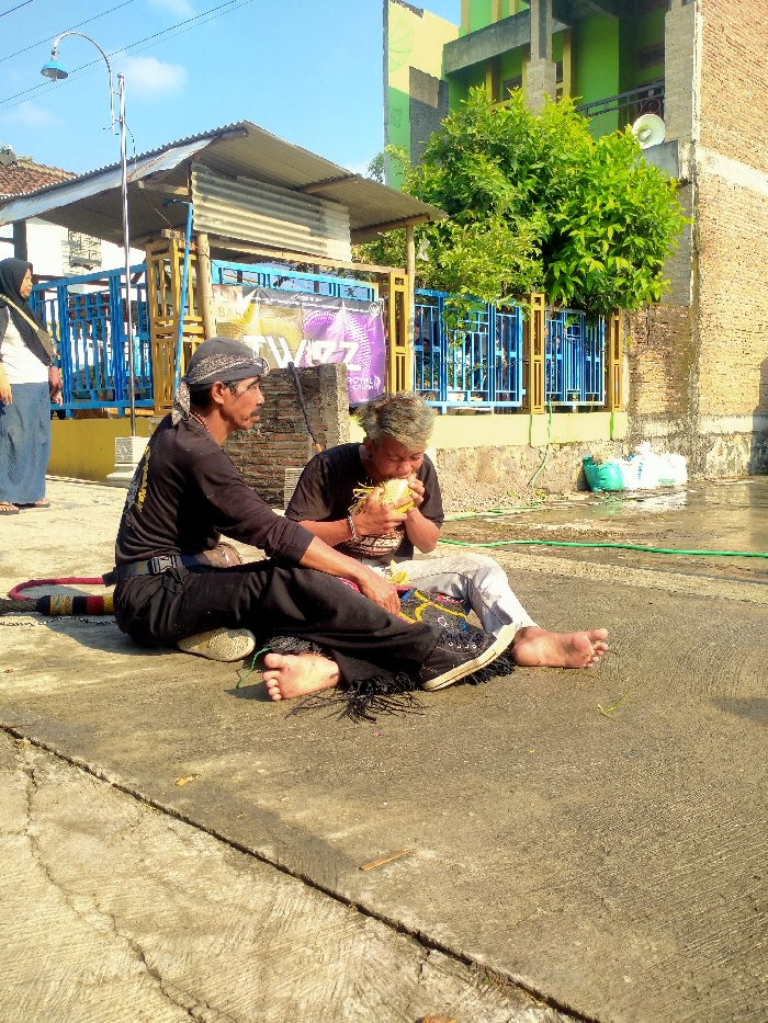The image depicts an outdoor scene in Sepat, Indonesia, under bright daylight. In the foreground, two adult males are seated on a cracked concrete surface. The man on the left wears a black long-sleeved shirt, black pants with fringed cuffs, and a black headscarf; his right foot is bare, while his left foot wears a dark shoe. He is positioned facing right. The man on the right, with light-colored hair, wears a dark short-sleeved shirt and light-colored pants. He is actively drinking from a green coconut, holding it to his mouth with both hands. His right foot is bare, and his left foot wears a dark high-top shoe with decorative elements. A multi-colored striped object lies on the concrete behind the left man.
In the midground, a structure with a corrugated metal roof supported by wooden posts is visible behind the seated men. Behind this, a blue metal fence or railing is partially obscured by a large purple banner displaying white text, including "TWOZ" and "ROYAL CRUSH." To the far left, an adult female wearing a light top, blue lower garment, and dark headscarf is partially visible standing. The background features a multi-story building on the right, characterized by exposed brick on the lower section and a green-painted upper section. Dense green foliage is present between the building and the fence. White bags and a green hose lie on the ground near the building's base. A tall lamp post with a curved arm and light fixture is positioned to the left of the central structure.