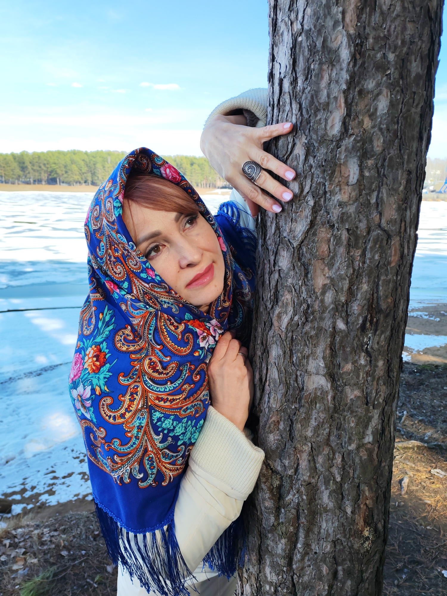 A woman with auburn hair and light skin poses outdoors next to a pine tree, wearing a blue patterned headscarf with fringe. She is dressed in a light-colored jacket or sweater. Her right hand is positioned on the tree with a large silver ring on her ring finger. Her left hand is also touching the tree. The background features a partially frozen body of water and a line of trees on the opposite shore, suggesting a lakeside or riverfront location. There is snow on the ground. The photo appears to be taken in the city of Chaikovsky, Russia.