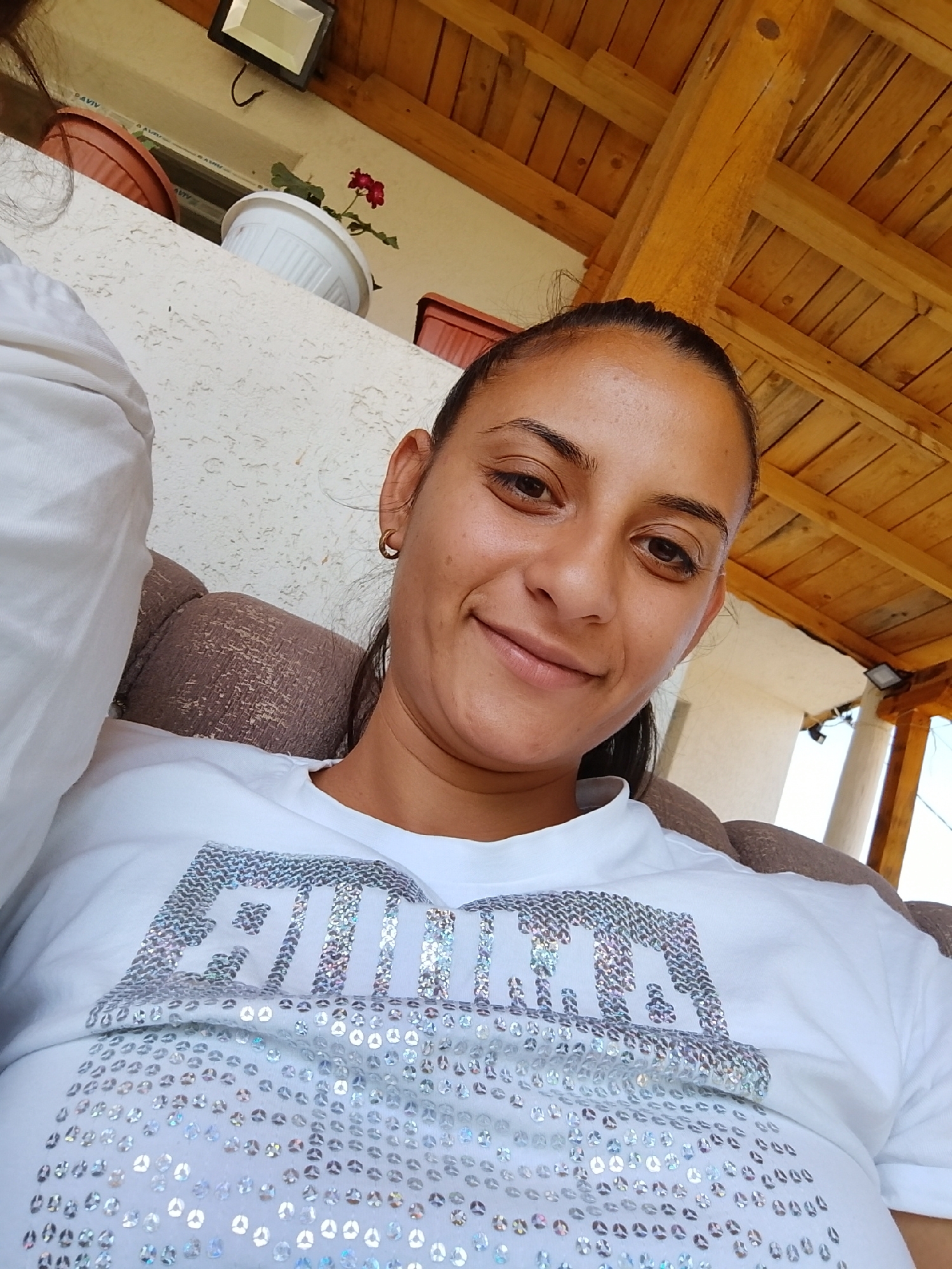 A woman wearing a white t-shirt with "PUMA" in large, silver sequins is sitting on a brown upholstered couch. She has dark hair pulled back and is looking directly at the camera with a slight smile. She is wearing a small gold hoop earring in her left ear. The background shows a textured white wall and a wooden ceiling with visible beams. To the left, there are two plant pots, one orange and one white, with a red flower in the white pot. A black outdoor light fixture is visible on the wall above the pots. The scene appears to be an outdoor covered area, possibly a patio or veranda, in Zimnitsa, Bulgaria.