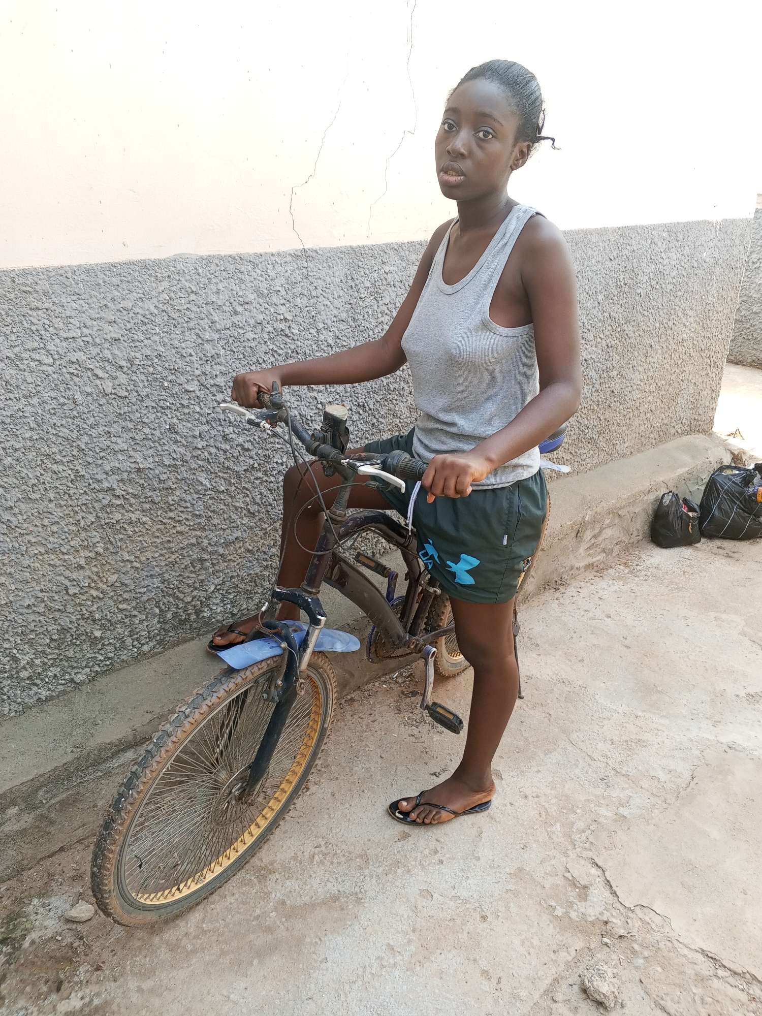 A young, dark-skinned woman stands next to a bicycle in an outdoor setting. She wears a grey tank top and green shorts with a blue logo. She has her left foot propped on the bicycle’s front tire and her hands on the handlebars. Her hair is pulled back. The bicycle is an older model mountain bike with a rusty frame and dirty tires. The background consists of a concrete wall with a rough texture. In the lower right corner are two black bags. The scene appears to be a residential area in a city in Nigeria. This is the result of an analysis based on the image content. There is no evidence to specifically indicate that the location is the city of Abuja.