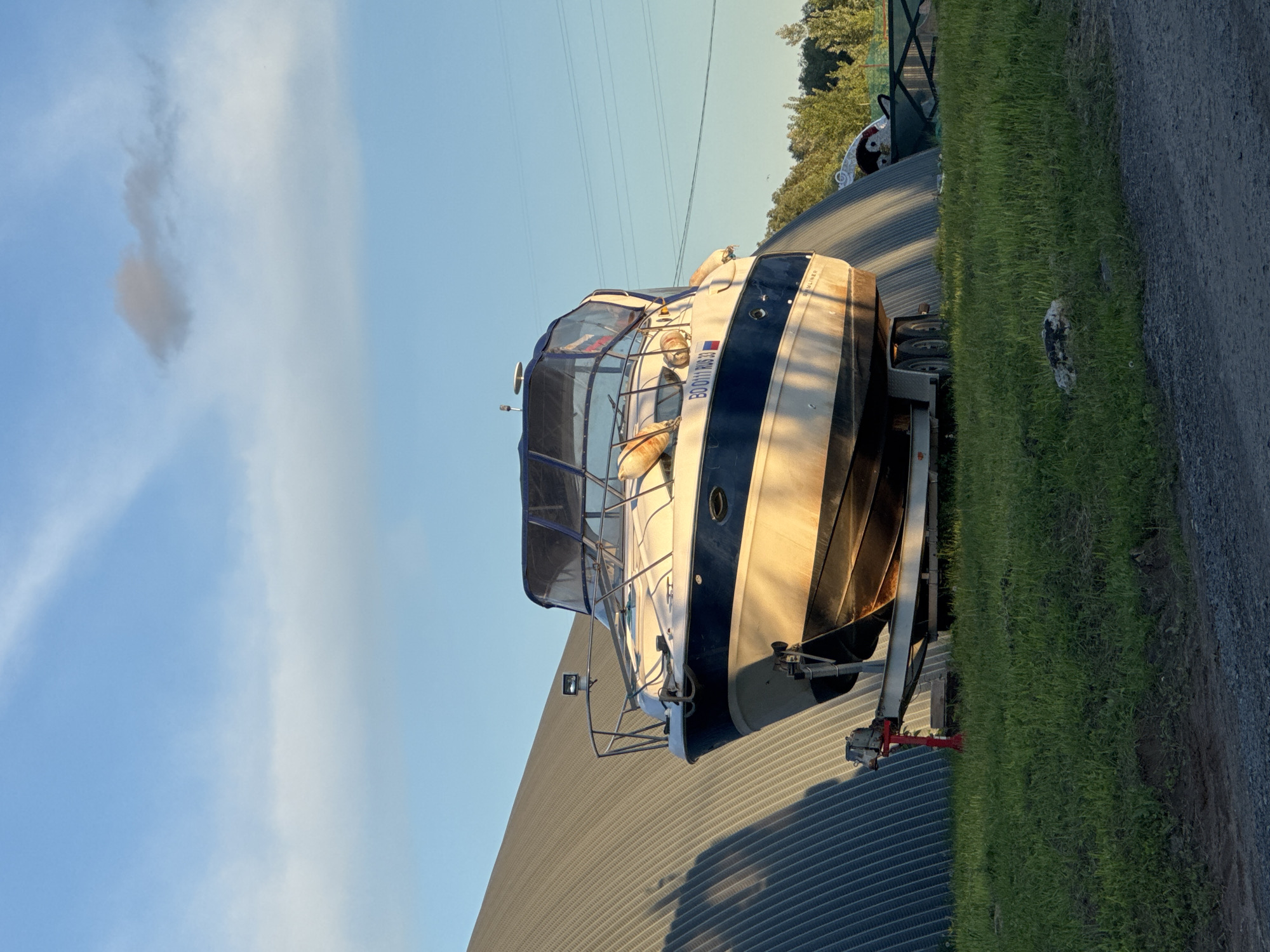 A medium-sized cabin cruiser boat sits on a trailer, positioned beside a low, corrugated metal building. Lush green grass grows along the right side of the frame, adjacent to a gravel or dirt path. The sky above is a gradient of blue, fading from a deeper hue to a lighter shade, with a few wispy clouds. Power lines stretch across the upper portion of the image. The location is Unknown, Unknown.