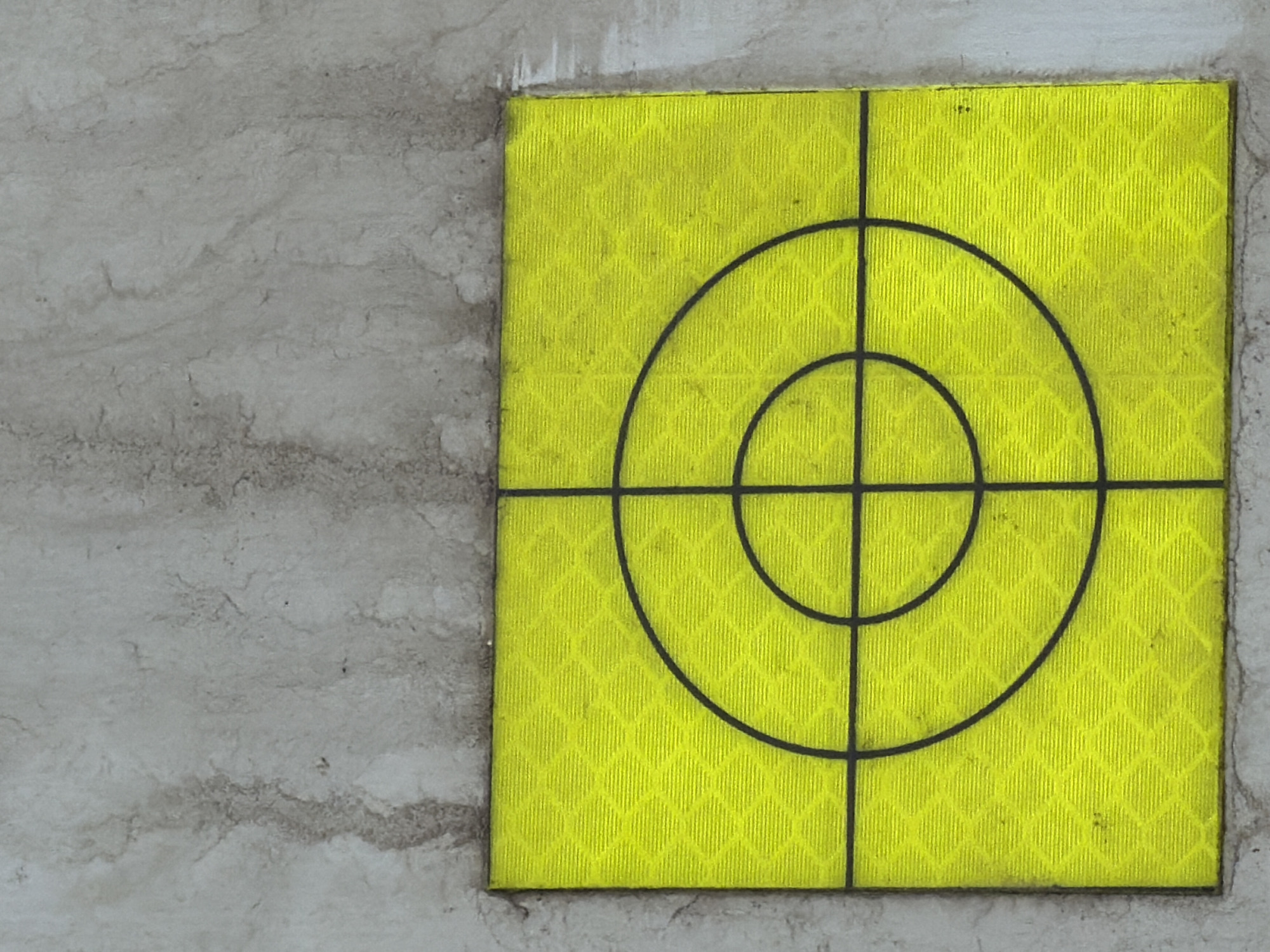 A close-up, overhead shot shows a bright yellow, reflective target with concentric circles and crosshairs, affixed to a light grey, textured concrete surface. The target occupies the bottom half of the image, while the top half is dominated by the weathered concrete. The concrete exhibits streaks of darker grime, suggesting it is an outdoor surface. The target itself has a subtle diamond-patterned texture beneath the black markings. There are no people, animals, or discernible signs of action or interaction. The scene appears to be static and located outdoors. The image is not associated with any specific location in Stróża, Poland.