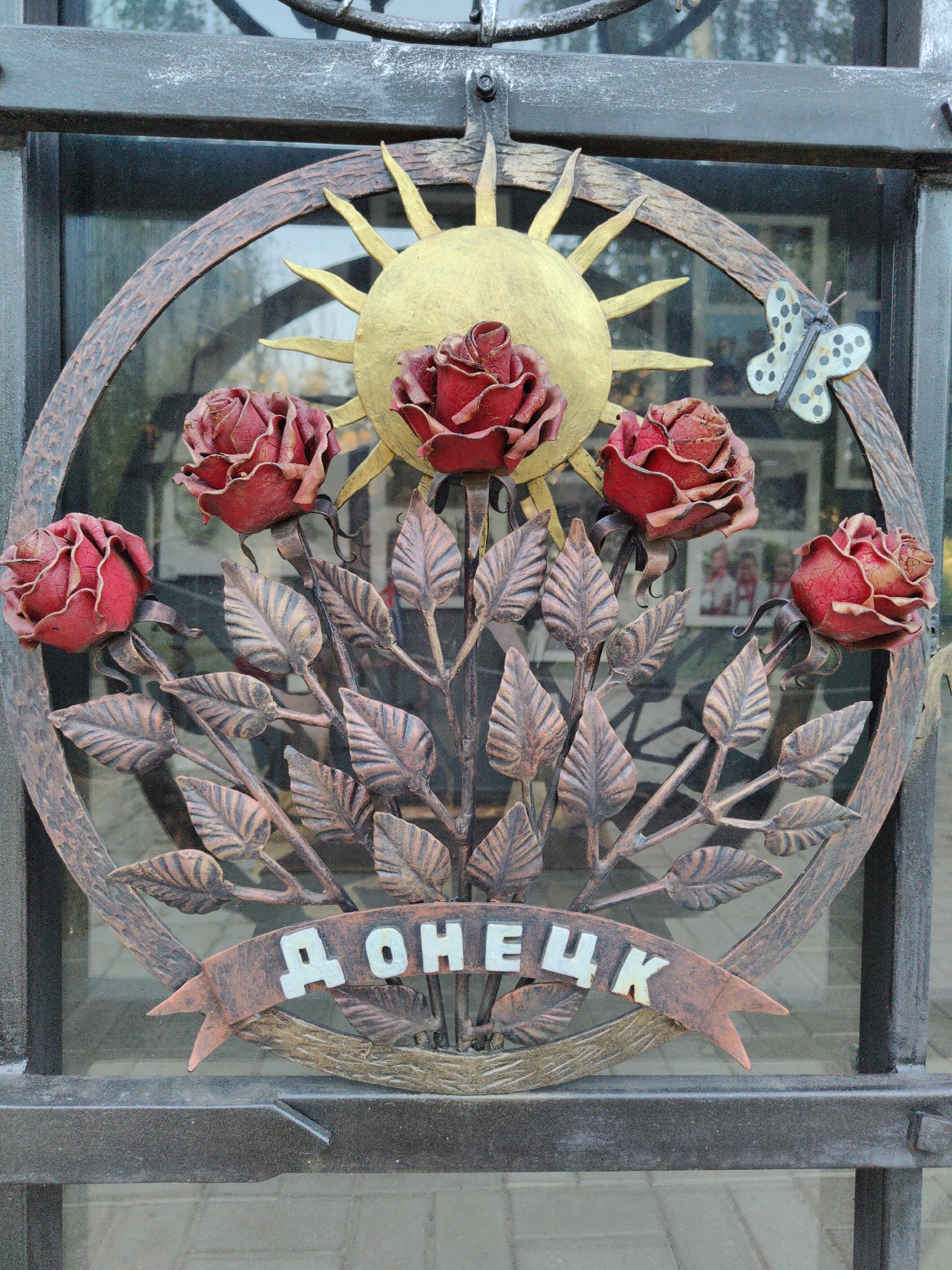 The image shows a decorative metal gate featuring a circular design with a sun and roses. The metalwork is ornate, with stylized red roses and leaves. A banner across the bottom of the circle displays the word "ДОНЕЦК" (Donetsk) in white, block letters. A small, white butterfly with black spots is attached to the right side of the circle. The gate appears to be in front of a window or glass pane, through which blurred images of people in frames can be seen. The overall impression is one of artistic craftsmanship, likely serving as an entrance or decorative element in Donetsk, Ukraine.