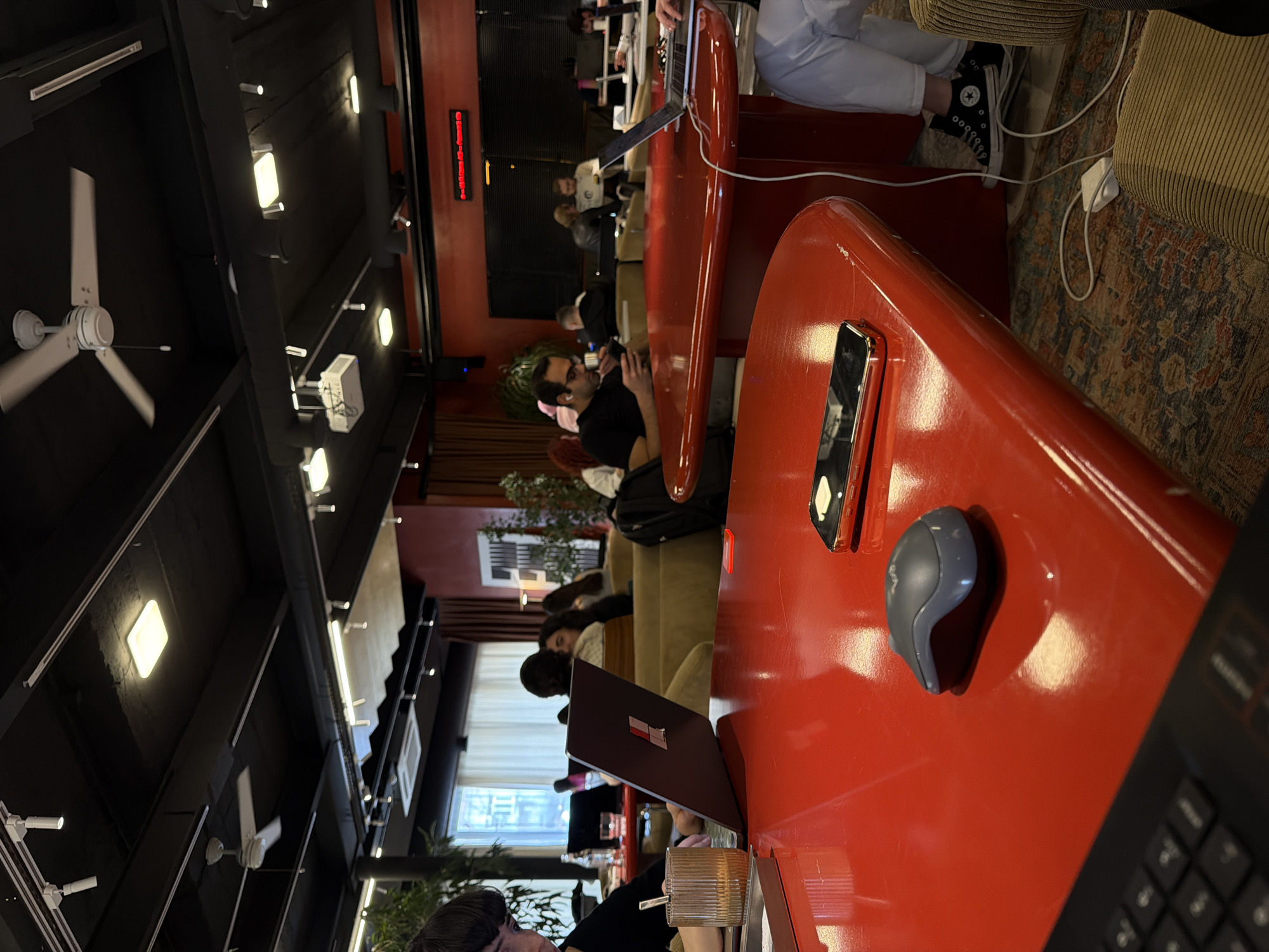 The image shows the interior of a co-working space in Yerevan, Armenia. The space has red tables with people working on laptops and using phones. Several people are seated on couches in the background, also working. The ceiling is dark with exposed lighting fixtures and a white ceiling fan. A digital display sign is visible on the wall. Several indoor plants can be seen throughout the space.