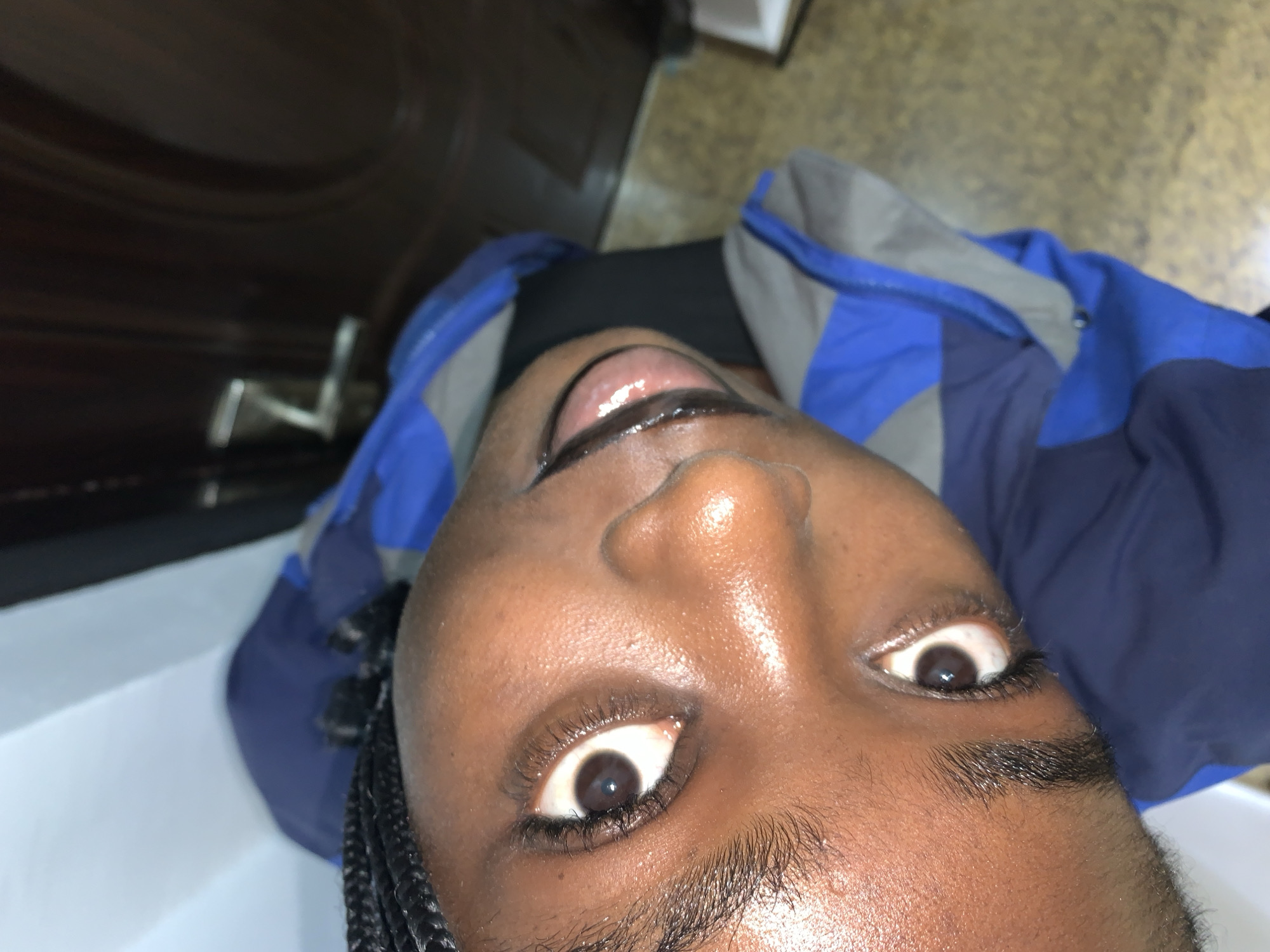 The image is a close-up, headshot of a young Black person wearing a blue and grey jacket. The person's face is tilted upwards, and their eyes are open and looking towards the camera. The person has dark braided hair. The background appears to be indoors, with a door visible on the right side of the frame. The floor is visible and appears to be tiled. The lighting is somewhat dim. The image does not provide any specific information about the location within Gura Top, Nigeria.