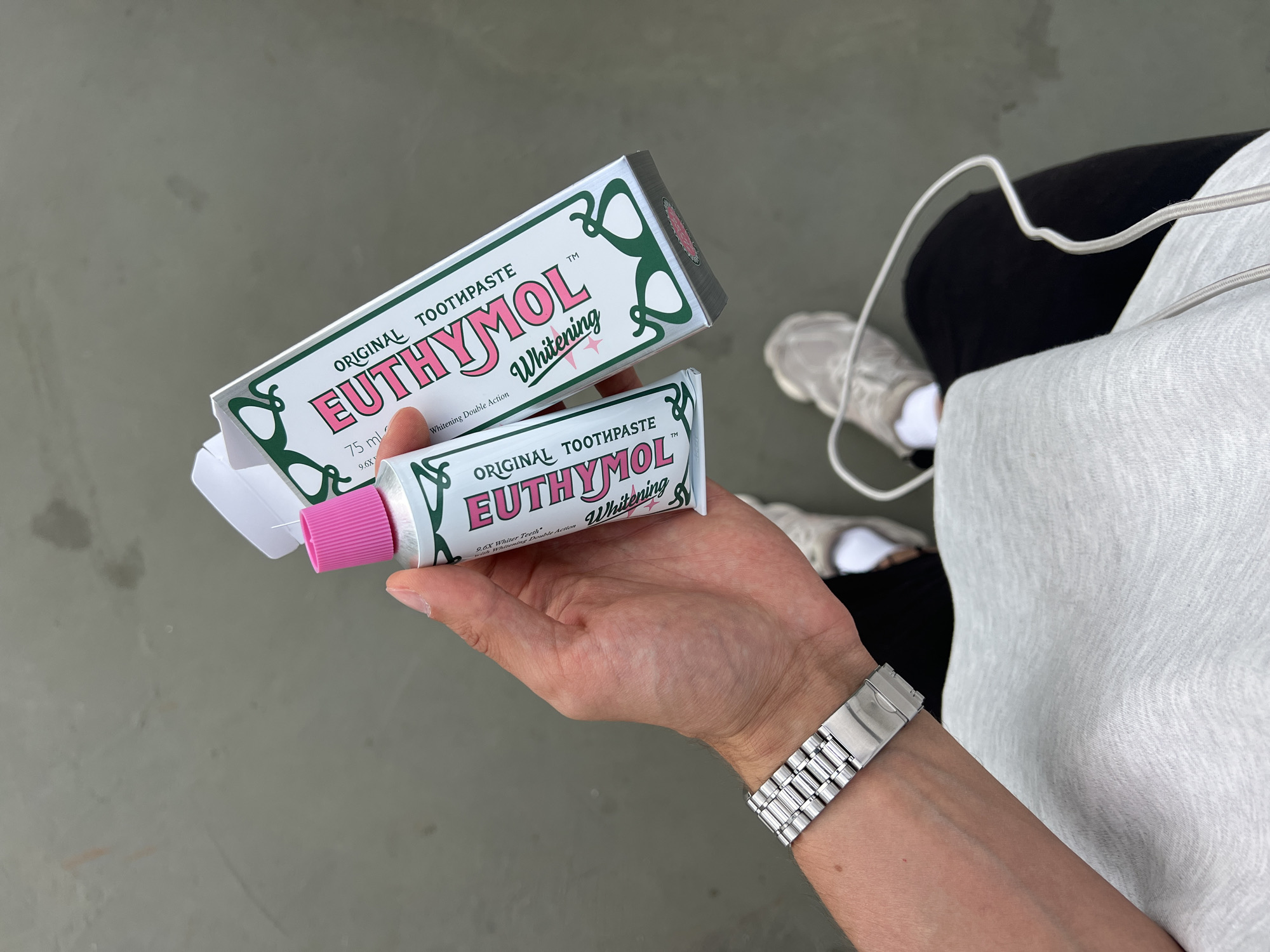 This media file features a close-up view of a person's hands holding a box and a tube of Euthymol Original Toothpaste. The toothpaste box is primarily white with green decorative accents and bold pink lettering that reads "EUTHYMOL." The tube, also white with similar branding, has a pink cap. The text "ORIGINAL TOOTHPASTE," "Whitening," and "Whitening Double Action" is visible on both the box and the tube. The person is wearing a silver wristwatch with a metal band. In the background, the lower body of another person is visible, wearing grey athletic shoes, black pants, and a light grey sweatshirt or hoodie. The scene appears to be outdoors, with a grey, textured ground surface. The lighting suggests it could be daytime. There are no strong indicators of weather conditions. The context provided indicates the location as Borowa, Poland. The overall impression is one of a casual, everyday moment, possibly shopping or preparing for a trip, with a focus on the iconic Euthymol toothpaste.