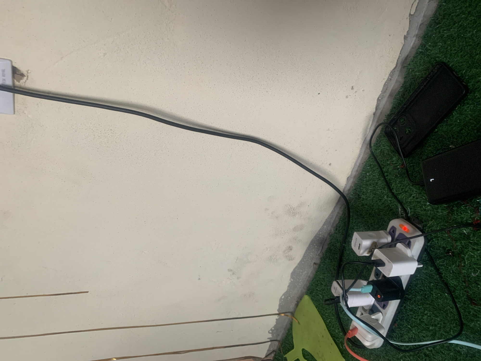 The image depicts a close-up view of a wall and a floor covered with artificial turf. A white electrical power strip is plugged into a wall socket via a black cable. The power strip is populated with several electrical adapters, some of which have indicator lights illuminated red, suggesting they are powering devices. Two black phone cases are lying on the artificial turf next to the power strip. The wall appears to be slightly stained or discolored in places. The scene is illuminated by artificial light, casting subtle shadows. The location is identified as Jauro Yinu, Nigeria. There are no people or animals visible in the image.