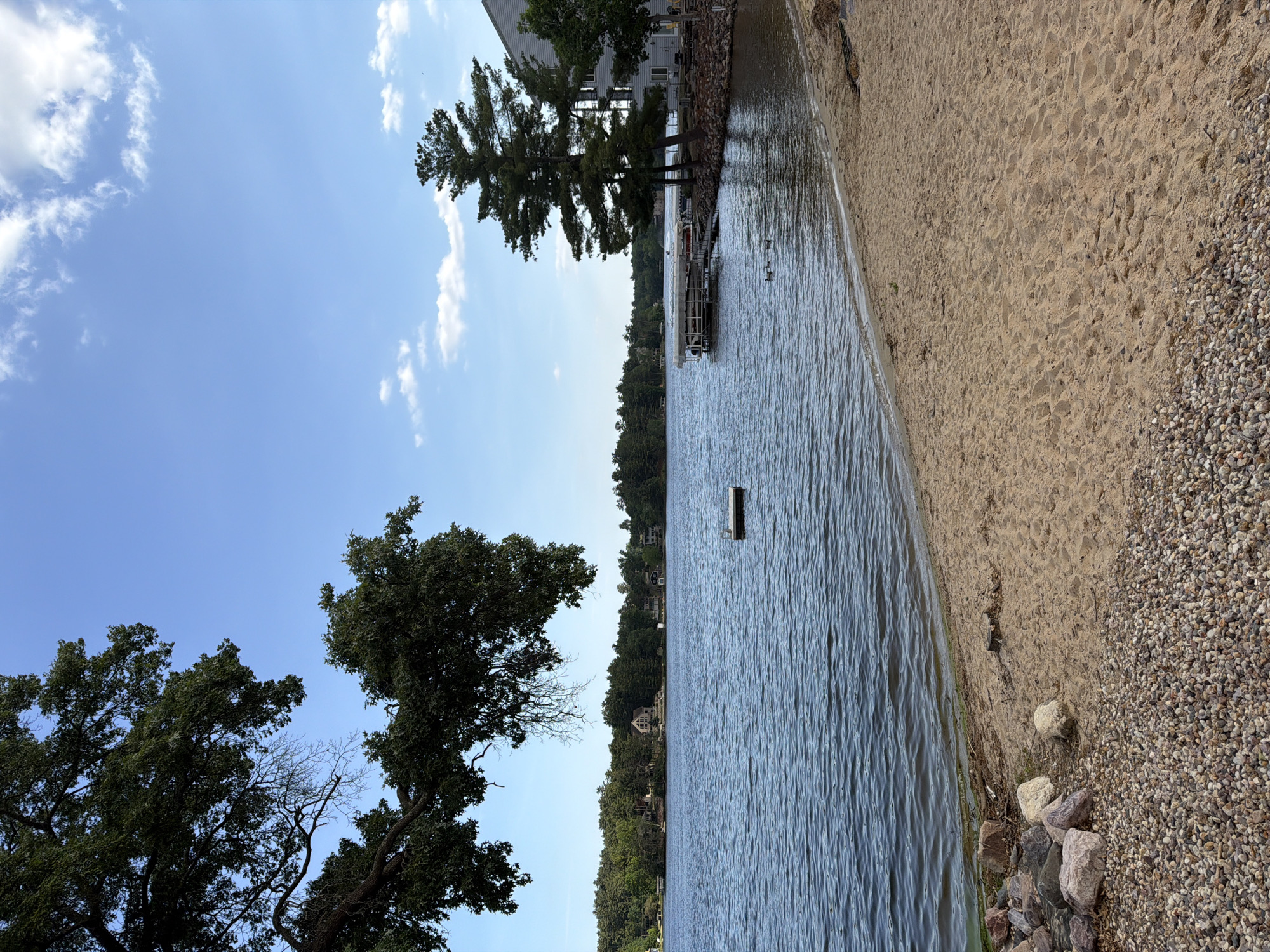 The image, when correctly oriented, captures a tranquil horizontal landscape view of a lake from a shoreline. The scene is dominated by a calm body of water under a bright blue sky with scattered white, fluffy clouds.
In the foreground, a strip of light brown sandy beach runs parallel to the water's edge, transitioning into a section of small, light-colored gravel and larger rocks further from the water. The lake itself shows gentle ripples on its surface, and a dark, rectangular buoy floats in the mid-ground.
To the left side of the frame, near the closer shoreline, a building and a dock with a covered boat (possibly a pontoon) are visible. Several tall pine trees stand near this structure. Further left and in the foreground, large, dark-leafed deciduous trees frame the view, with one showing a few bare branches contrasting with the full foliage.
Across the lake, a distant shoreline is heavily wooded with lush green trees, interspersed with several houses, suggesting a residential area nestled along the lake. The overall impression is one of a clear and sunny day, likely morning or afternoon, at Lake Delton, United States of America, conveying a peaceful and serene atmosphere. No people or specific activities are depicted. No text is visible in the image.
