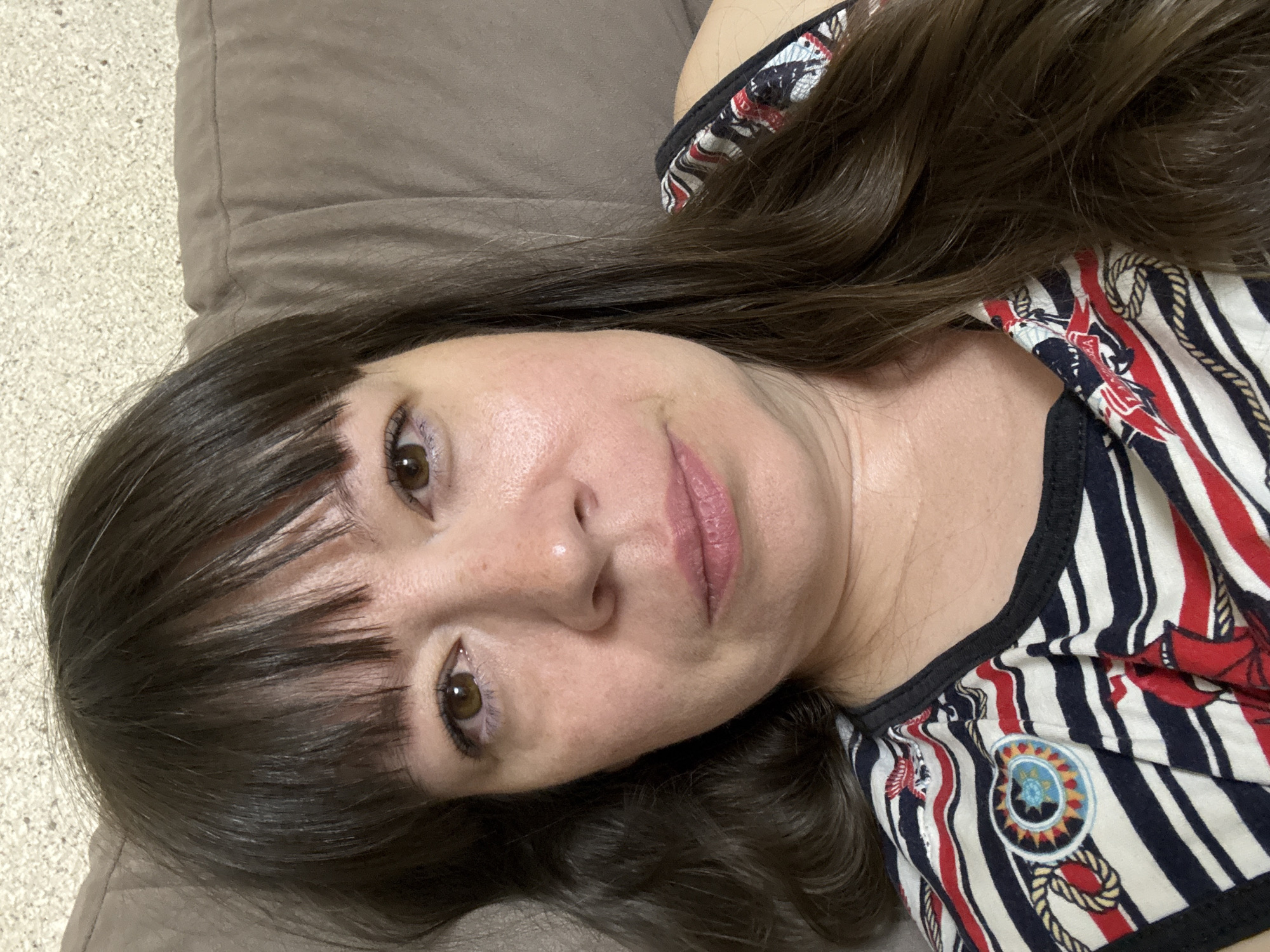 A medium shot shows a woman with dark brown hair and brown eyes from the chest up, looking directly at the camera. She is wearing a black, white, red, and blue striped shirt with nautical-themed graphics. The woman is lying down on a brown couch, with the back of the couch visible behind her head. The scene appears to be indoors, possibly in a living room. The location in the city of Ветлужский, Russia is not determinable from the image.