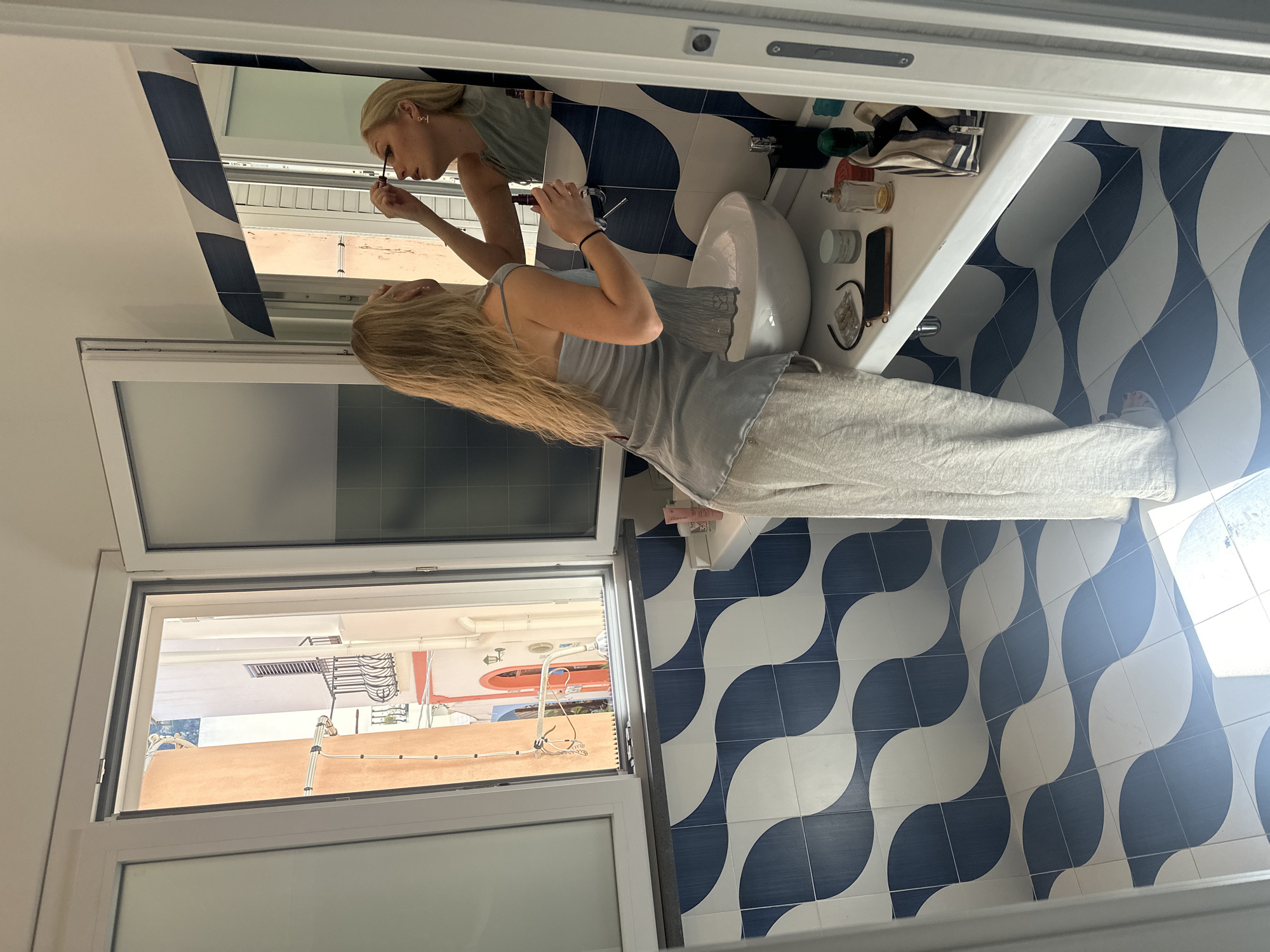 A blonde woman is depicted applying mascara while looking into a mirror in a well-lit bathroom or vanity area. She is wearing a light grey tank top and light-colored, possibly linen, pants. Her reflection is clearly visible in the mirror, showing her focused on her task.
The area around the sink is white, with a small, modern white sink bowl. On the countertop next to the sink, various personal items are arranged, including several perfume bottles, a blue and white striped makeup bag, small jars or containers of cosmetics, a dark object resembling a mobile phone, and a pair of eyeglasses or sunglasses. A small clear container with capsules or pills is also present.
The bathroom features a distinctive floor and wall tile pattern composed of interlocking blue and white wavy shapes. To the woman's left, an open window offers a view of the outside. Through the window, parts of buildings with terracotta-colored walls and white accents are visible, along with some external piping or cables, suggesting a typical Mediterranean street or alleyway. The bright light entering through the window indicates it is daytime. A white towel or piece of fabric is partially visible on a surface towards the bottom right of the frame. No visible text is present in the image. The scene captures a personal moment of grooming in a stylish setting, consistent with a location like Lipari, Italy.