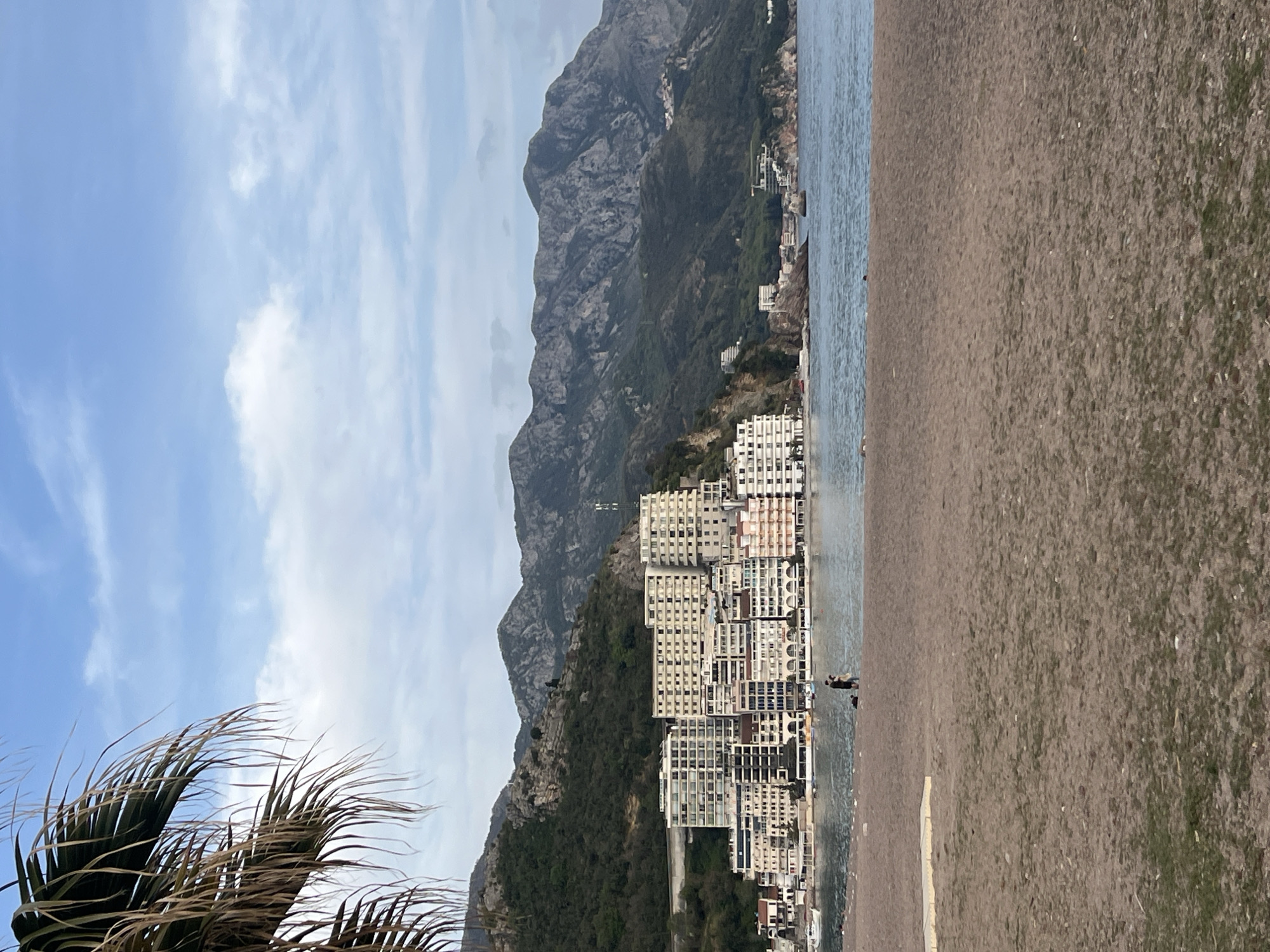 The image shows Rafailovići, Montenegro. It is a bright, overcast day. In the foreground is a beach, and beyond it is the Adriatic Sea. The right side of the image is dominated by a tan-colored beach of small rocks. In the middle ground, tall, densely packed, high-rise hotels and apartment buildings stand close to the shoreline. Rocky mountains with vegetation rise behind the buildings. In the top left, the sky is visible with a mix of blue sky and white clouds. A palm tree partially obscures the lower left corner. Two people are standing on the beach near the shoreline in the middle of the frame.