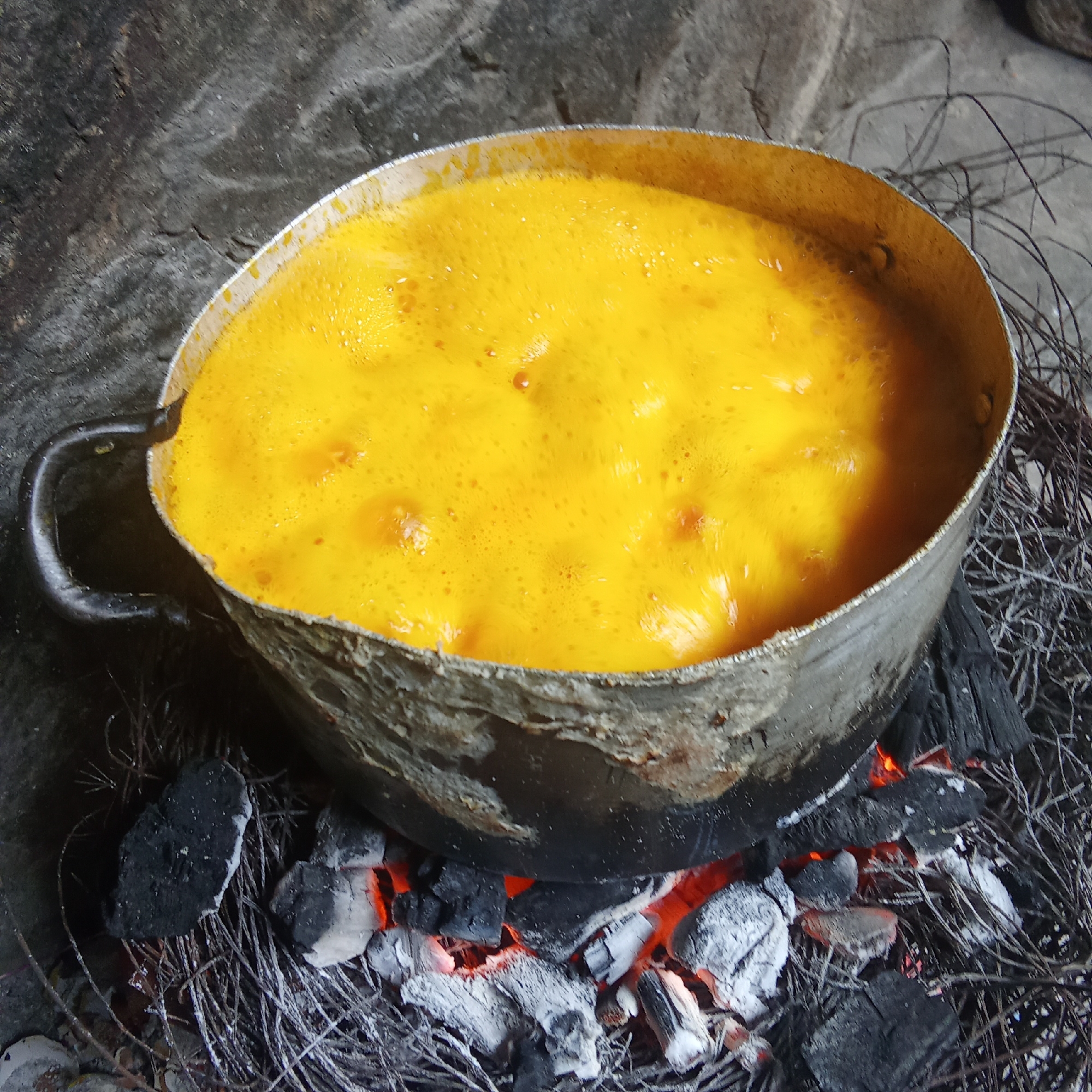 A close-up shot depicts a rustic cooking scene featuring a well-used, metallic pot brimming with a vibrant orange-yellow liquid. The liquid is actively boiling, with a frothy, bubbly surface indicating intense heat. The pot, showing signs of wear and discoloration, has a simple handle on its left side. It is positioned directly over a makeshift fire, consisting of glowing red and white charcoal embers mixed with ash. These hot coals are resting on a bed of dark, dry, fibrous material, possibly twigs, dried grass, or straw, suggesting a traditional or outdoor cooking method. The background is dark and indistinct, featuring a rough, textured surface, likely ground or a simple hearth. There are no people visible, nor any discernible text. The image focuses on the process of cooking, highlighting the rich color of the bubbling contents against the dark, earthy tones of the heat source and surroundings.