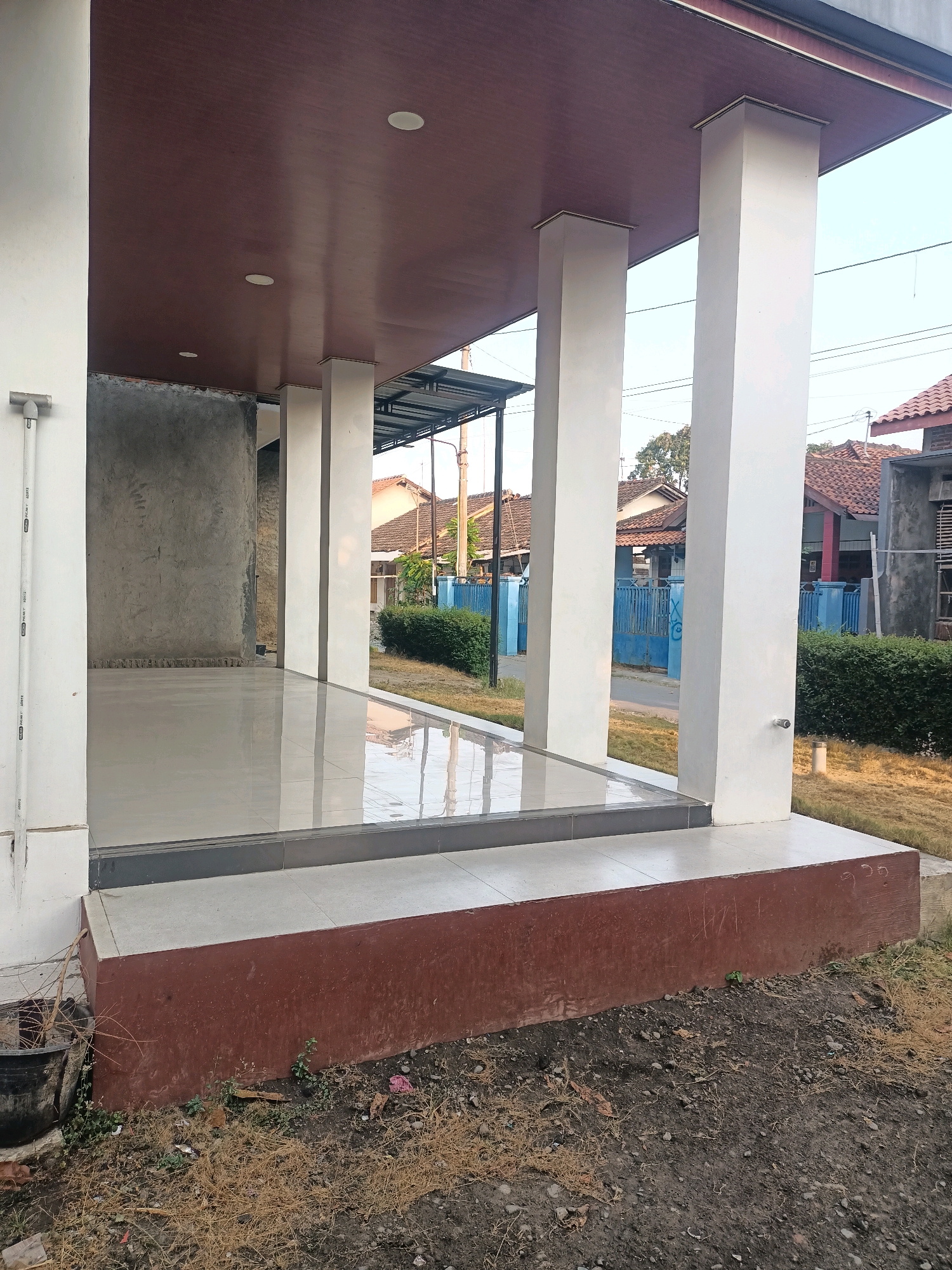 The image shows an outdoor view of a building's exterior, possibly a veranda or a walkway. The structure features white pillars supporting a brown ceiling with recessed circular lights. The floor of the veranda is tiled in a light gray color and is highly reflective. To the left, an unfinished concrete wall is visible. The area in front of the veranda is covered in dry grass and dirt. Behind the building, houses with red tile roofs and blue fences are visible, suggesting a residential area. The scene is set during the daytime, under a clear sky. The location is identified as Tembok Luwung, Indonesia.