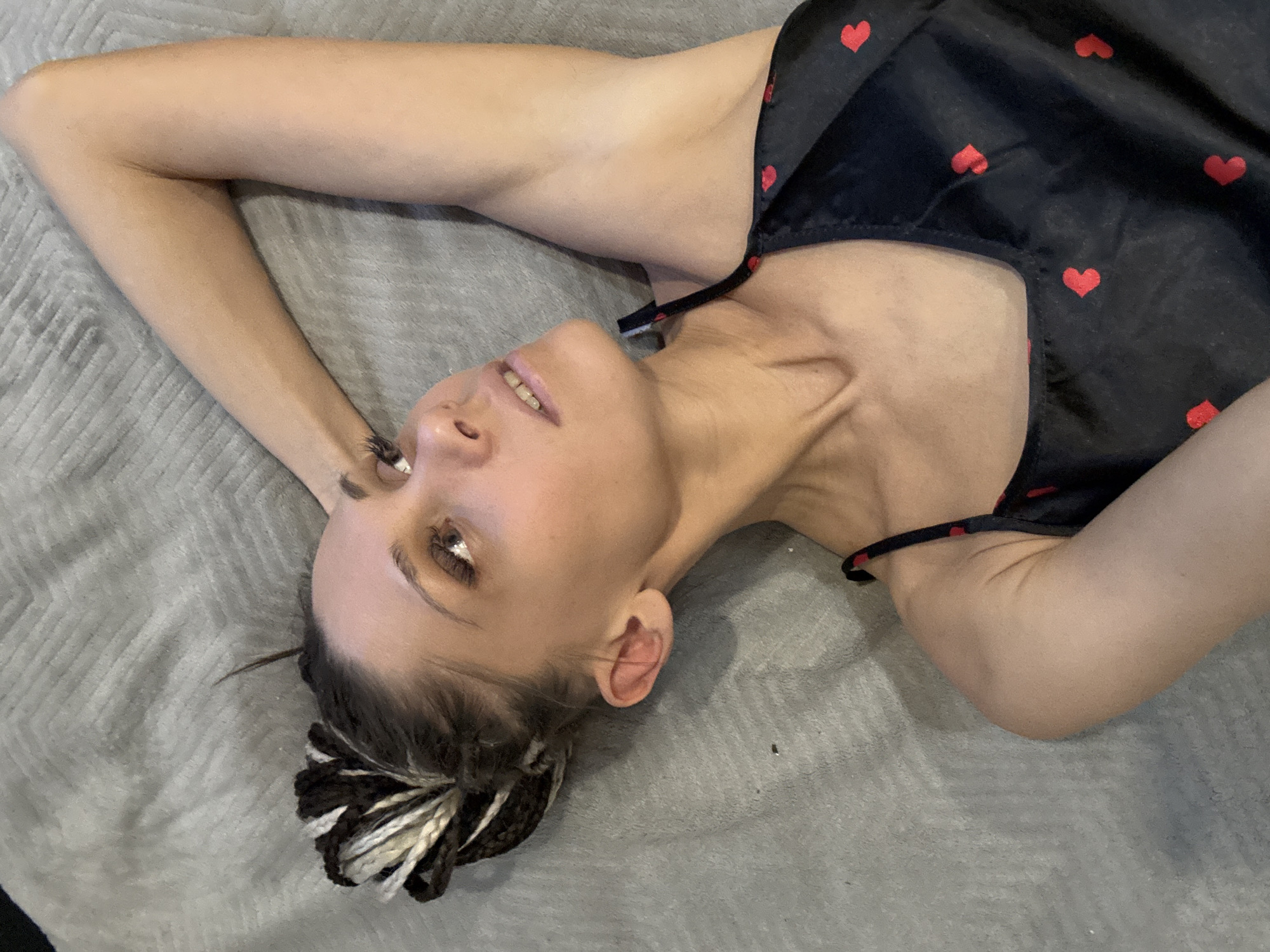 A woman is lying on a gray surface. She is wearing a black top with red hearts. Her arms are above her head, and she is looking up. Her hair is styled in black and white braids. The location is unspecified, and there is no evidence to suggest the location is Yaroslavl', Russia.