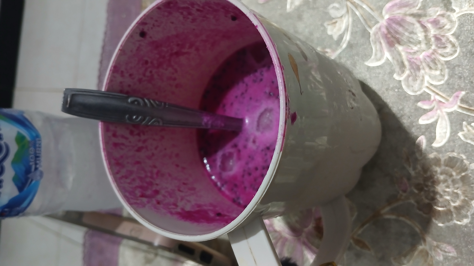 A close-up, eye-level shot shows a white mug filled with a vibrant pink liquid, likely a dragon fruit smoothie, with a metal spoon partially submerged in it. The mug's interior is stained pink, suggesting it has been used for this drink before. The mug has a white handle and rests on a table with a floral pattern. In the background, a partially visible bottle of water with a blue and white label stands behind the mug. The lighting is dim, creating shadows on the table and highlighting the texture of the drink. The overall scene appears to be set in a domestic environment, possibly a kitchen or dining area. Without further context, the exact location within Sampang, Indonesia cannot be determined from the image alone.