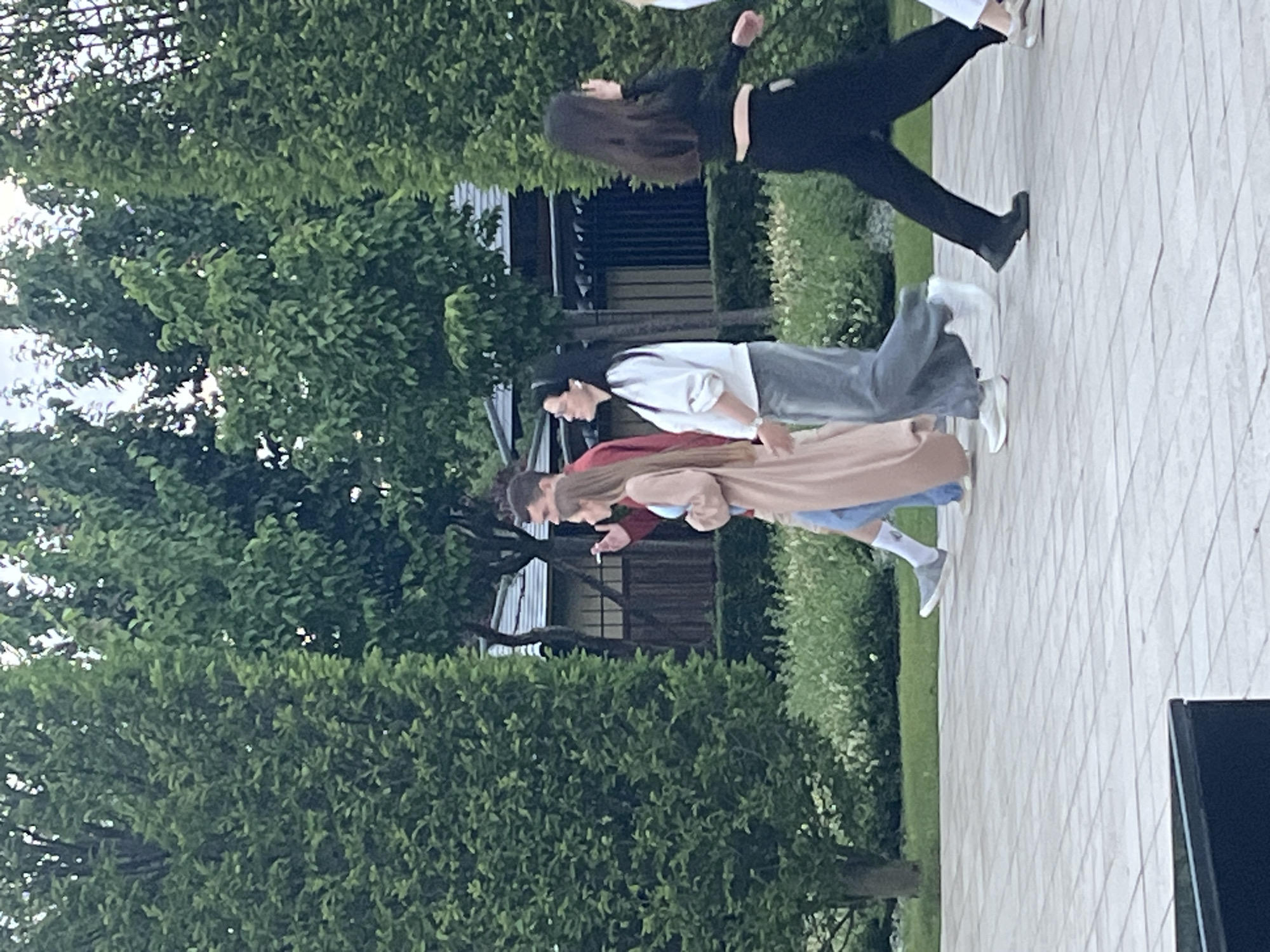 This outdoor image, taken in Krasnodar, Russia, features a group of at least four people standing on a light-colored paved area, backed by lush green trees and foliage.
The subjects are arranged in a loose line. On the left, a person with long dark hair, dressed in a black long-sleeved top and black pants, is captured in a dynamic, mid-movement pose, with one arm raised and a leg extended, possibly dancing or in mid-stride. Next to them stands an individual wearing a white long-sleeved top, grey pants, and white earbuds. Their dark hair is pulled back, and they appear to be looking forward.
Further to the right, a person wearing a red top beneath a light brown or beige long jacket is visible, holding what appears to be a cigarette in their right hand. Partially obscured behind them, the legs and feet of a fourth person are seen, clad in light blue jeans, white socks, and grey sneakers.
The setting is characterized by a wide expanse of dense green trees and bushes forming a natural backdrop. Behind the subjects and within the foliage, a dark-colored building or structure with horizontal wooden slats or siding is partially visible. The ground beneath the people is a paved surface, made of light-colored tiles or stones arranged in a diagonal grid pattern. To the right of the paved area, a strip of green grass is discernible, and a dark, rectangular object, possibly outdoor furniture, is positioned on the far right.
The lighting is natural and bright, suggesting daytime, possibly a late afternoon or overcast day, with no strong shadows. The overall impression is one of a casual gathering or outdoor activity in a pleasant, verdant environment. No text is visible in the image.