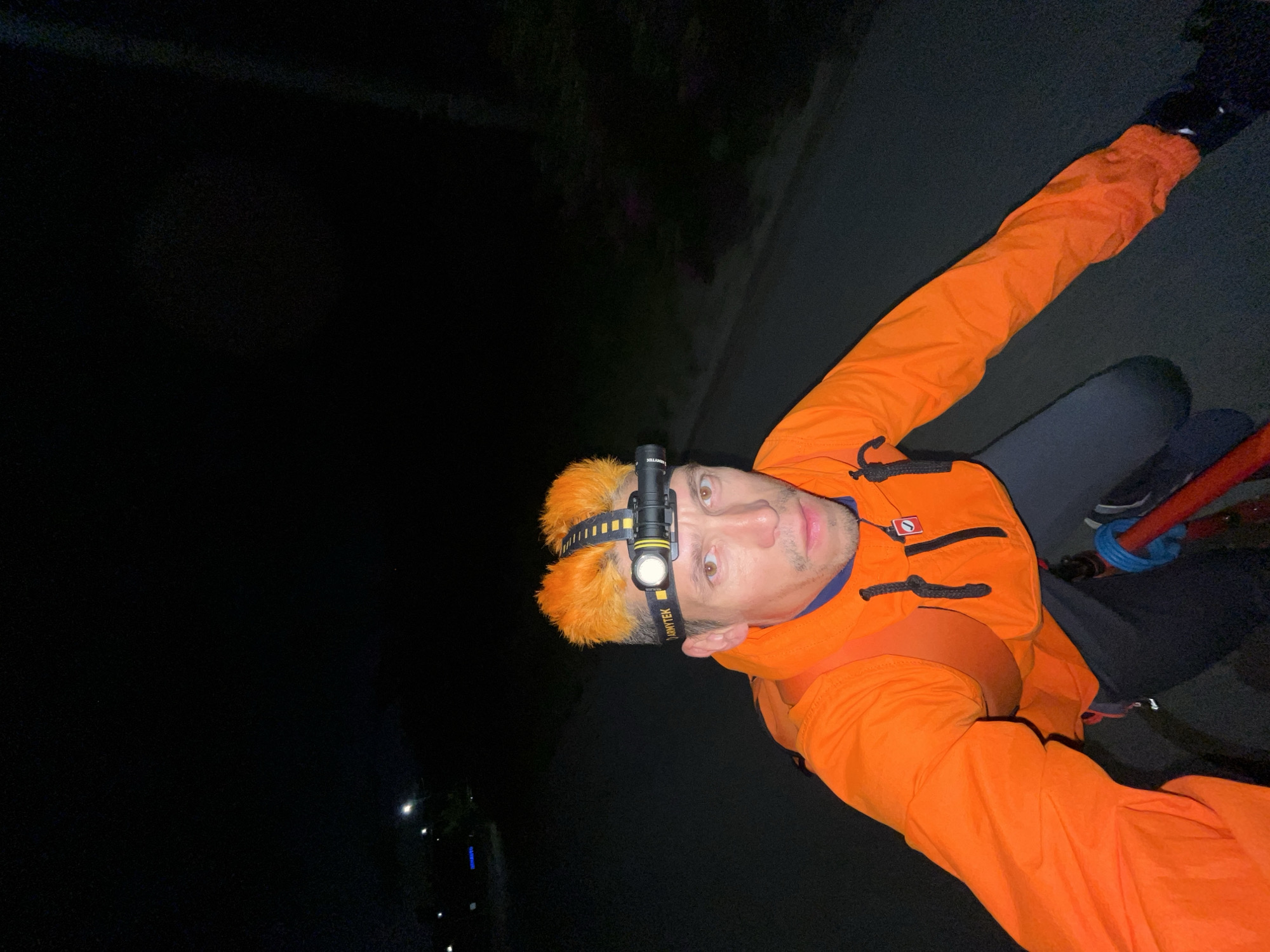 An individual with brightly dyed orange hair is riding a bicycle at night. They are wearing an orange hooded jacket and a headlamp with a white LED light. The individual is looking at the camera. The background is dark, indicating nighttime, with a few distant lights visible. The scene appears to be on a paved road. The general location is identified as Chaikovsky, Russia.