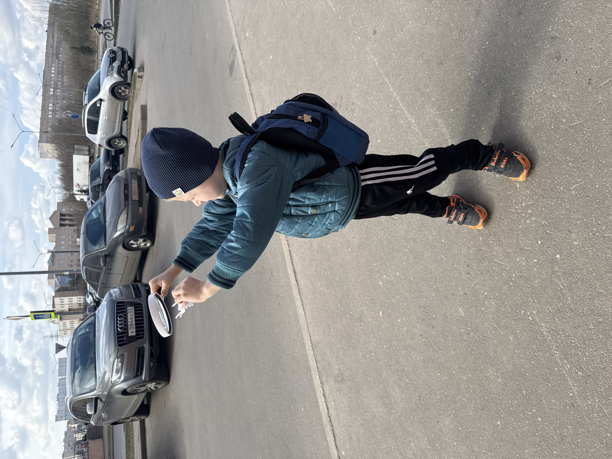 A child stands on an asphalt surface in Veliky Novgorod, Russia. The child wears a navy knit cap, a teal quilted jacket, black Adidas-striped pants, and black and orange sneakers, with a blue backpack. The child holds a ring light and a white cloth. Several parked cars line the curb, including a gray Audi with a visible license plate: K084KO 53. A building is visible in the background. The weather appears sunny with clouds.