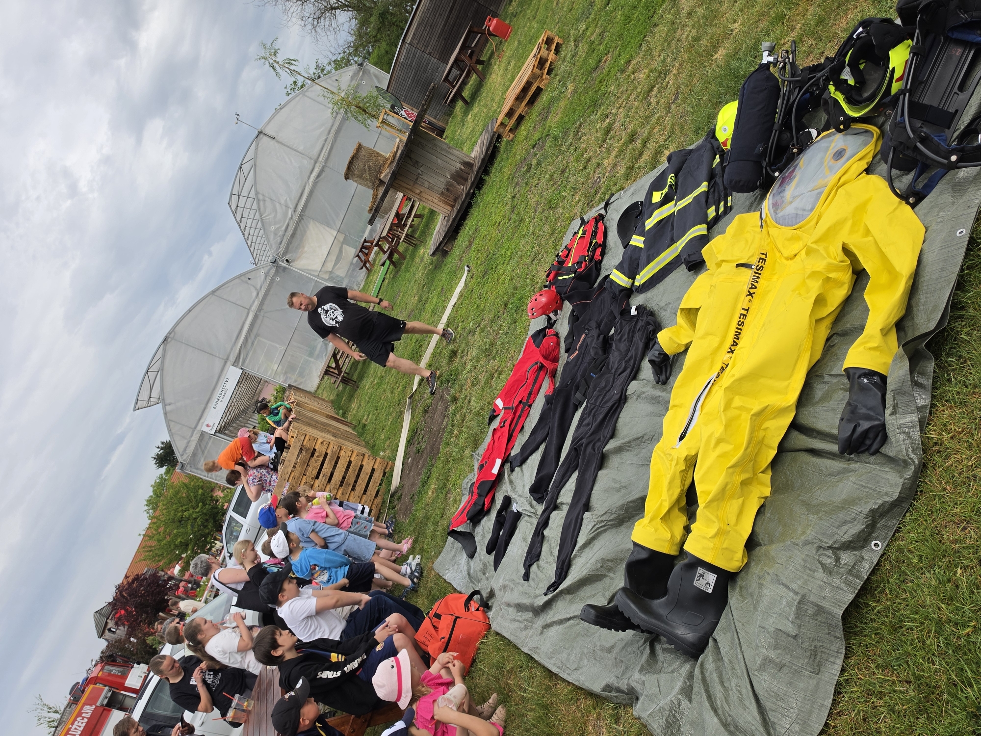The image shows a scene in Mlčechvosty, Czechia. A group of people, mostly children, are seated on a bench, facing a presentation of firefighting equipment. The equipment is laid out on a green tarp on the grass. It includes a yellow hazmat suit, black and red fire-resistant clothing, helmets, and oxygen tanks. A man in a black t-shirt and shorts is standing near the equipment, possibly giving the presentation. In the background, there are greenhouses and wooden structures. A red fire truck is visible on the left side of the frame. The sky is cloudy.