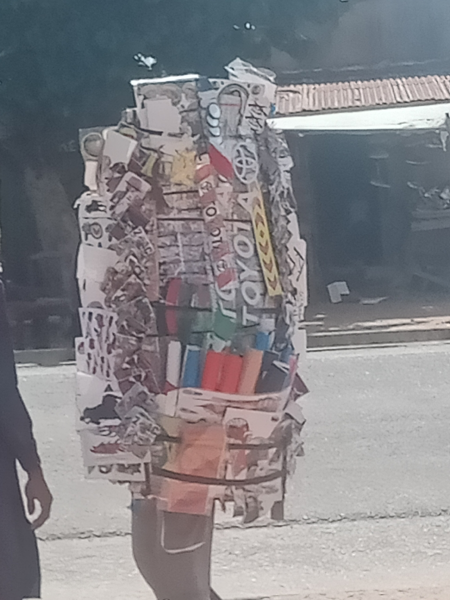 The image shows a person in Jalingo, Nigeria, carrying a large, stacked load of items. The load appears to be composed of various packaged goods, possibly snacks or other retail items, wrapped in plastic and secured with straps. Some of the packaging features colorful graphics and text, with the word "TOYOTA" clearly visible on a yellow and red sign. The person is standing on a paved surface, likely a road or sidewalk, with a building and trees visible in the background. Another person, partially visible on the left side of the image, is facing away from the camera. The overall scene suggests a street vendor or someone transporting goods for sale or delivery.