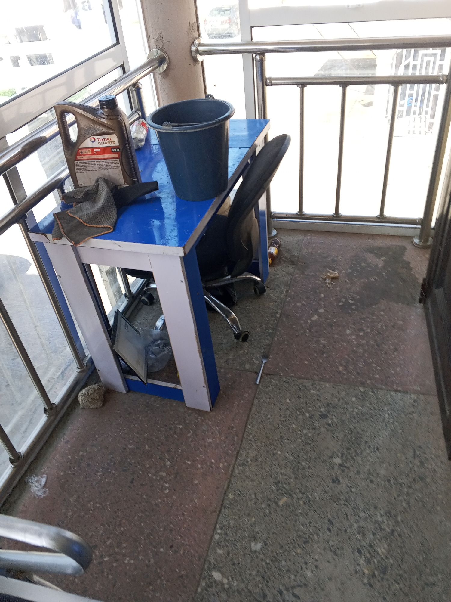 This image depicts an outdoor, possibly semi-enclosed, space with a view through a metal railing. In the foreground, a blue and white table holds a black plastic bucket and a bottle of "TOTAL QUARTZ" engine oil, along with a folded gray cloth. To the right of the table, a black office chair is partially visible, positioned on a speckled floor composed of red and gray concrete tiles. A small, silver object resembling a fork lies on the floor near the chair. A light-colored rock is situated near the base of the table. The background shows a bright, possibly sunlit area through the railing, with glimpses of buildings and possibly a vehicle. The overall scene suggests a somewhat cluttered or utilitarian area, perhaps a service or storage space, possibly located in Jalingo, Nigeria, given the context provided, though the image itself does not contain explicit identifying markers for this location.