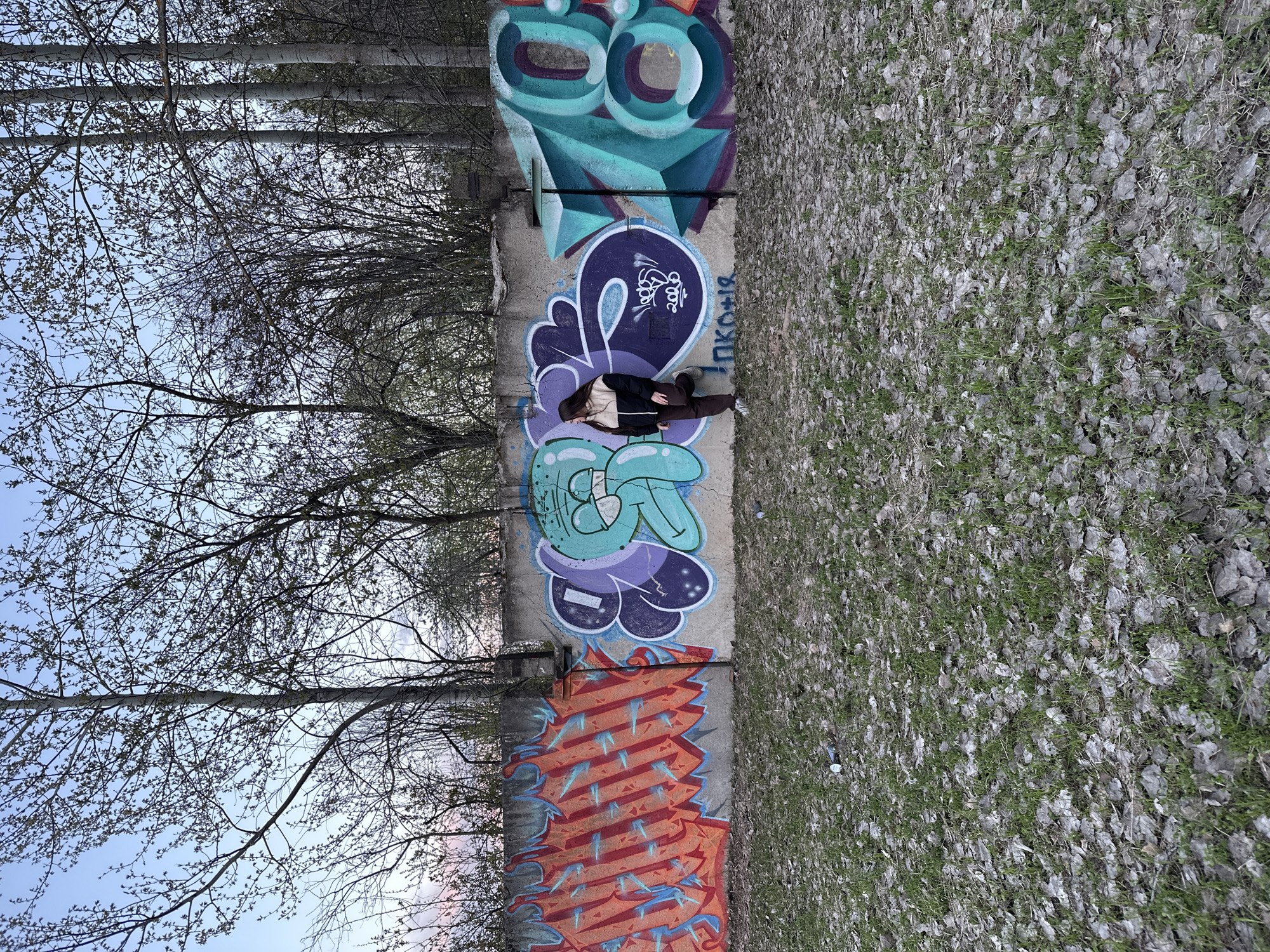 A person is lying on a concrete wall covered with graffiti in an outdoor environment. The graffiti includes various colors and shapes, with some resembling letters and numbers. The person is wearing a black jacket, brown pants, and light-colored sneakers. The wall is flanked by trees on one side and a ground covered in leaves and grass on the other. The location is Chaikovsky, Russia.