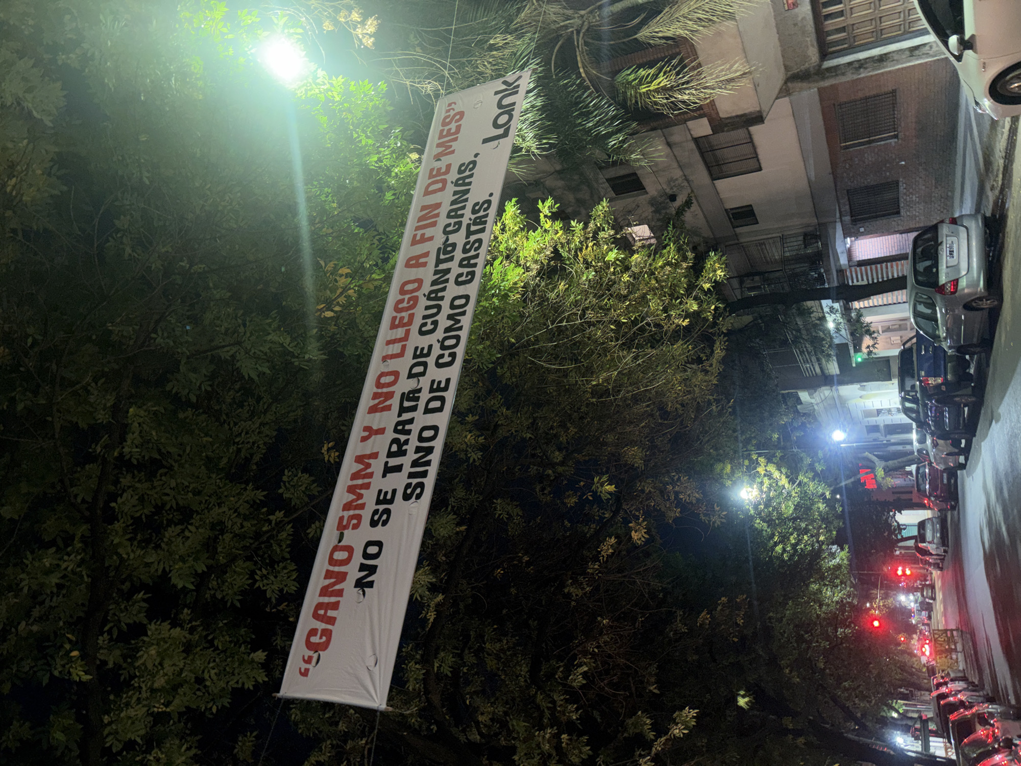 The image is a street scene at night, likely in Buenos Aires, Argentina, suggested by the Spanish text on a banner. The banner reads "GANO-5MM Y NO LLEGO A FIN DE MES" "NO SE TRATA DE CUÁNTO GANÁS. SINO DE CÓMO GASTÁS. Lonk". It is hung vertically between trees. The street is illuminated by streetlights. Cars are parked along the right side of the street, and traffic lights are visible in the distance. Buildings line the street behind the trees.