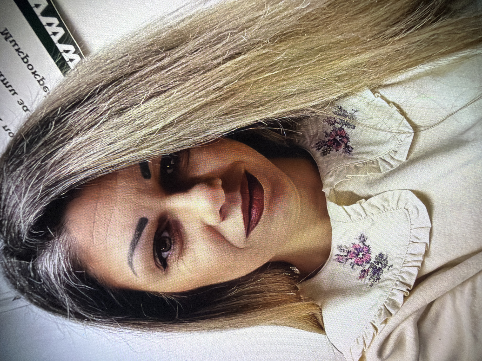 A woman with long blonde and grey hair is facing forward, positioned at an angle to the camera. She has dark eyebrows, dark eyes, and dark red lipstick. She is wearing a light-colored top with a ruffled collar embroidered with floral patterns. The image appears to be taken indoors. The text "MM" is visible in the top left corner, along with a word "mkbokbs" and "30 ur" at the bottom. The location in the city of Dryanovo, Bulgaria is not evident in the image.