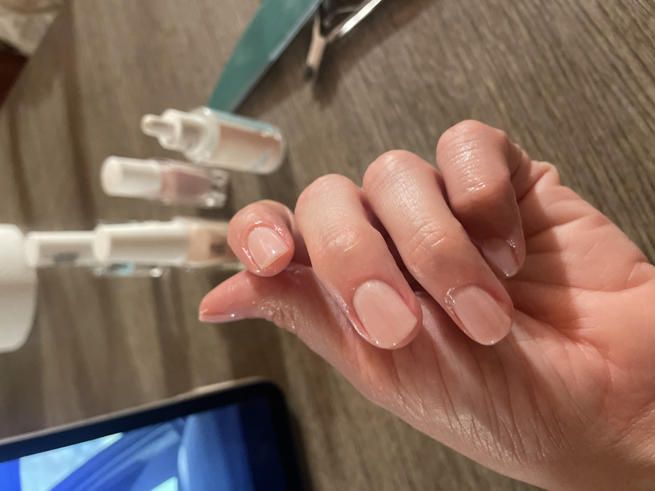 A close-up, slightly elevated shot captures a person's left hand resting on a wooden surface in Monsummano Terme, Italy. The hand displays five fingernails, each coated with a translucent, light pink or nude-colored nail polish. The skin on the fingers and palm exhibits a distinct sheen, indicative of recent application of moisturizer or cuticle oil. In the blurred background, positioned on the same wooden surface, are various nail care items: four small bottles with white caps, likely containing nail polishes or treatments, including one with a dropper applicator. To the right, a teal-colored nail file and a metal cuticle nipper are present. A dark electronic tablet or smartphone, with a blue-toned screen, is partially visible on the far left of the frame.