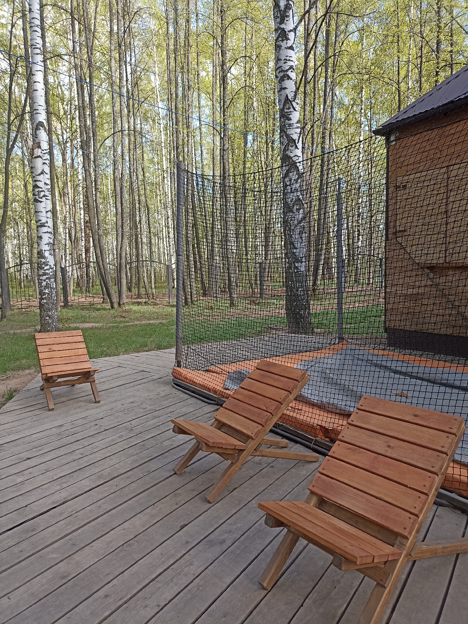 The image shows an outdoor recreational area in a forest-like setting. The scene is dominated by a wooden deck with three wooden chairs placed on it. Two chairs are positioned closer to the viewer, while the third one is further away. In the background, there is a safety net surrounding what appears to be a trampoline. Beyond the net, a dense collection of birch trees fills the landscape, suggesting a forest or wooded area. To the right of the frame, part of a wooden structure with a dark roof is visible, possibly a shed or small building. The environment suggests a place for relaxation or outdoor activities within a natural setting. The location is identified as Unknown, Unknown.