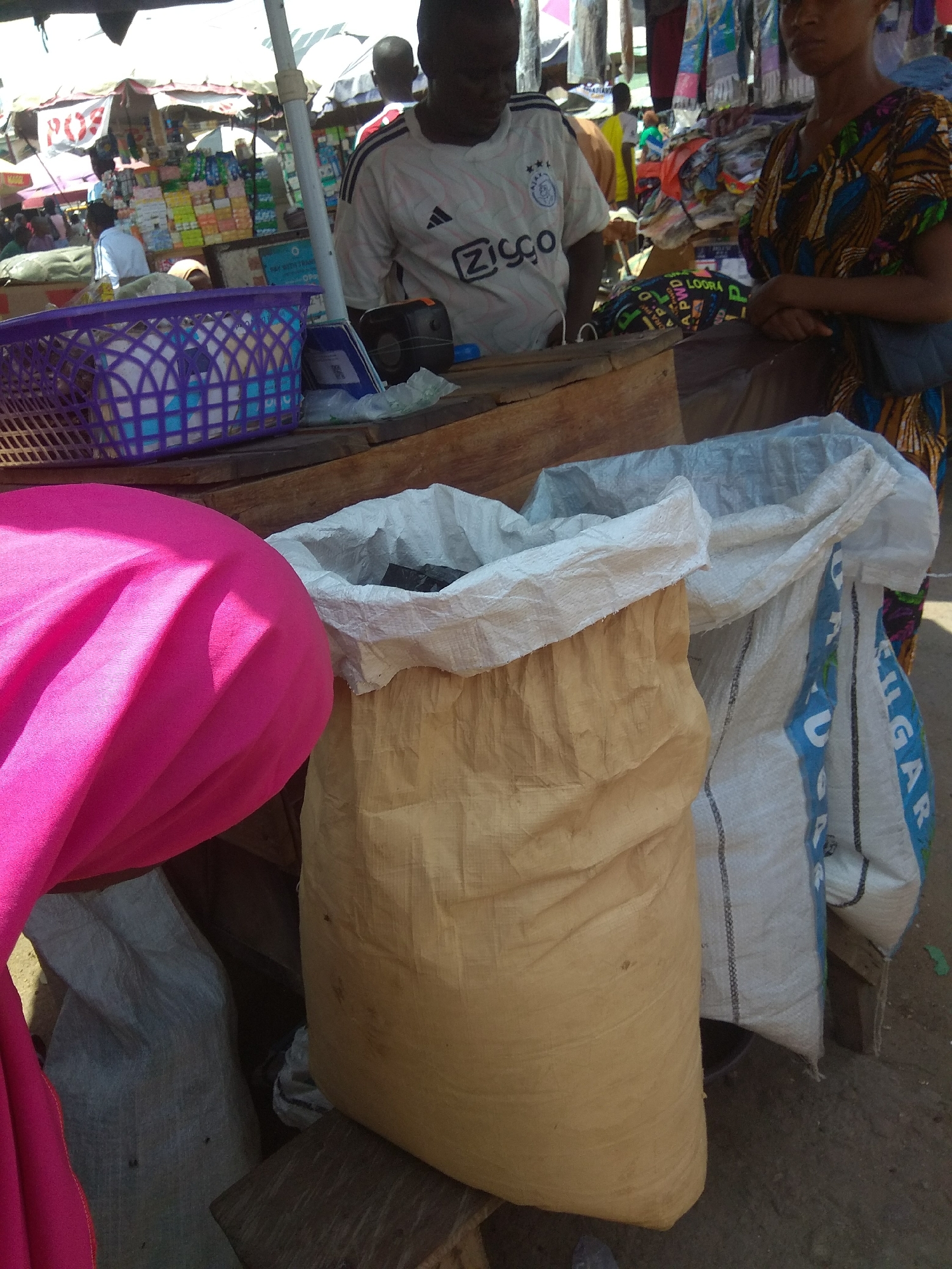 The image depicts a market scene in Jalingo, Nigeria. In the foreground, a large, tan-colored burlap sack is prominently displayed, partially obscuring a bright pink fabric in the lower left corner. To the right of the burlap sack is a white woven plastic bag with blue lettering. Behind these bags, a wooden surface is visible, holding a purple basket and a black electronic device.
In the background, several people are partially visible. A person wearing a white Adidas shirt with black stripes stands behind the wooden surface. To their right, a person in a colorful patterned dress is also present. Further in the background, market stalls are visible, filled with various goods, suggesting a bustling commercial environment. The overall setting appears to be outdoors, under a canopy or partial shade, consistent with a market.